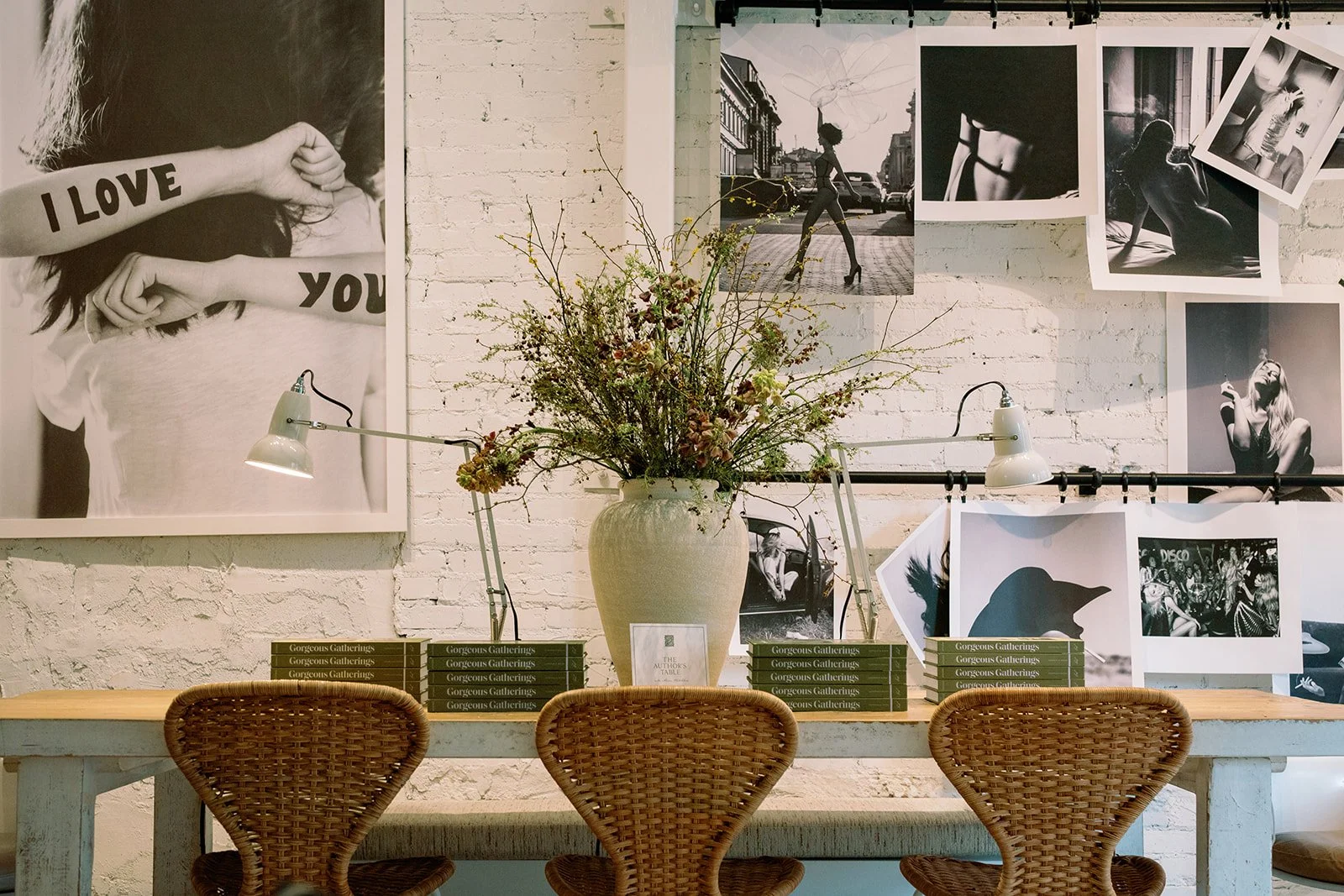 Interior wall decorated with black and white photographs, a large vase with flowers, and a stack of books, with three wicker chairs in front.