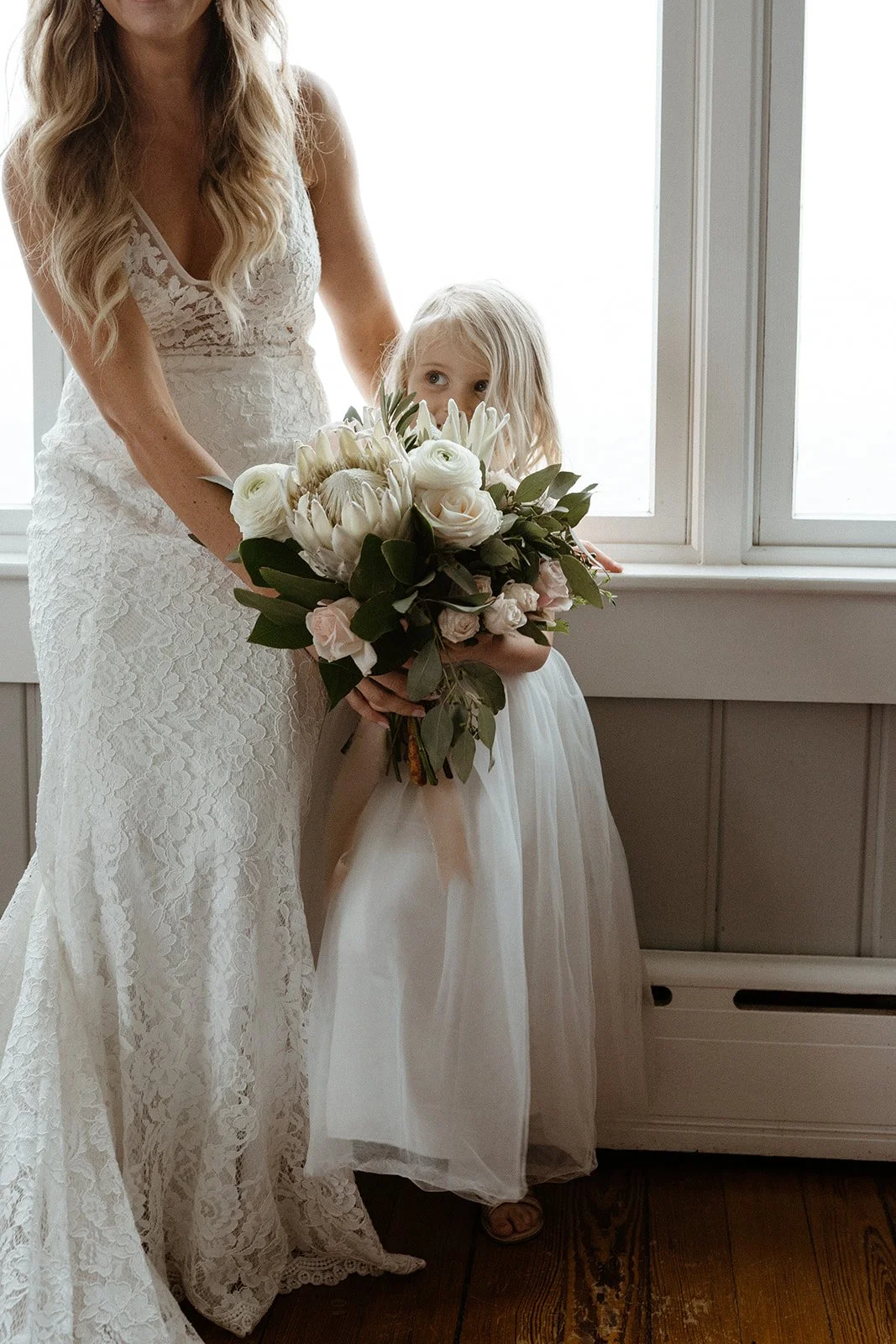 A woman in a white lace dress holding a bouquet of white flowers with a young girl in a white dress standing beside her near a window.