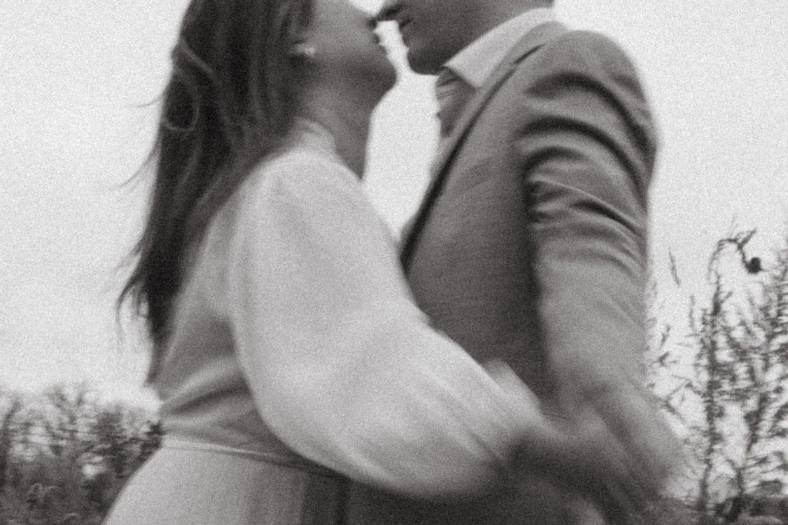 A black and white photo of a couple kissing outdoors, with trees in the background.