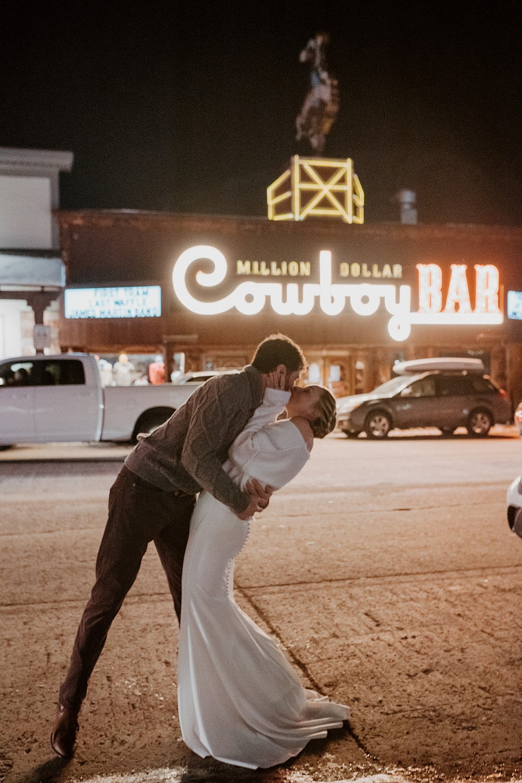 A man and woman sharing a kiss outside a bar at night, with neon sign reading 'Million Dollar Cowboy Bar' in the background.