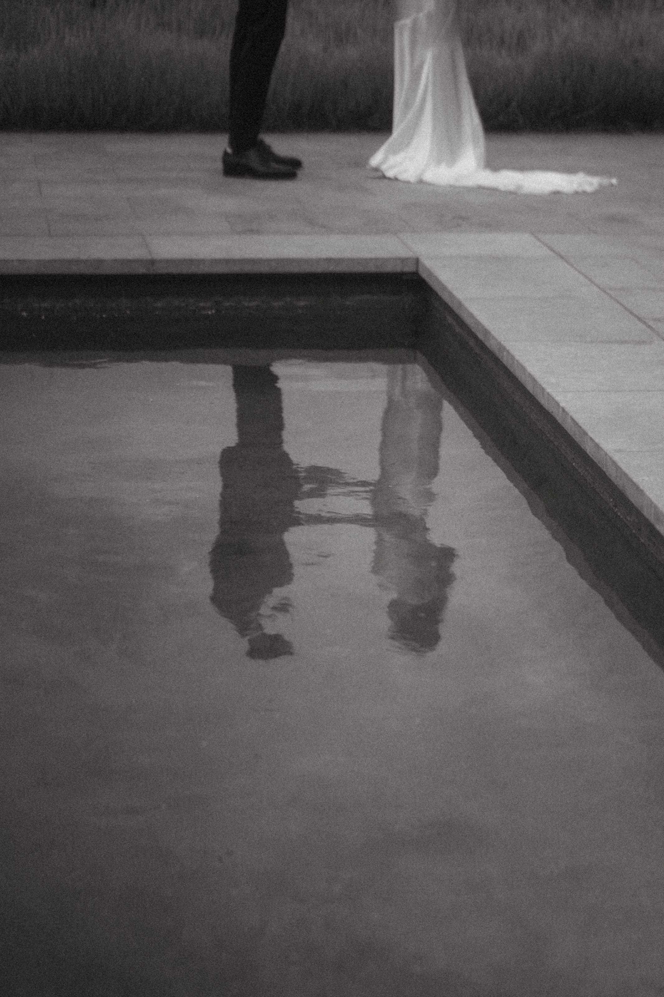 A couple holding hands by a pool, with their reflection visible in the water.