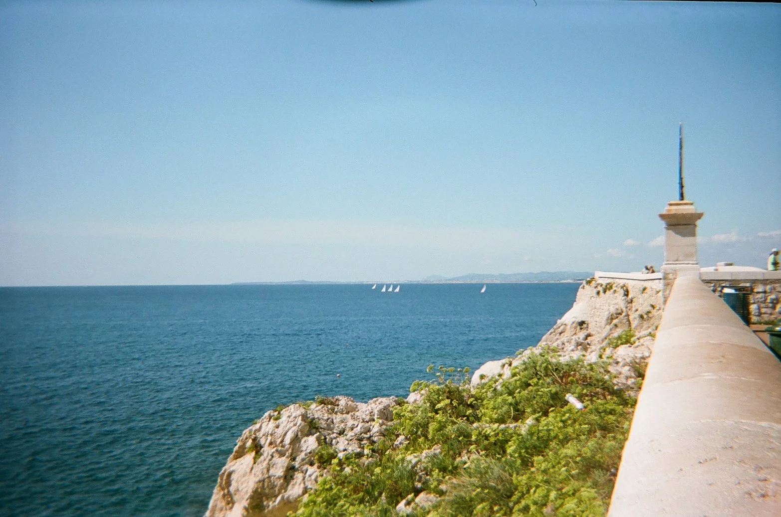 A coastal scene with a concrete pier or promenade extending along the rocky shoreline, a stone wall with a small monument at its end, and a distant view of the ocean with sailboats on the horizon under a clear blue sky.