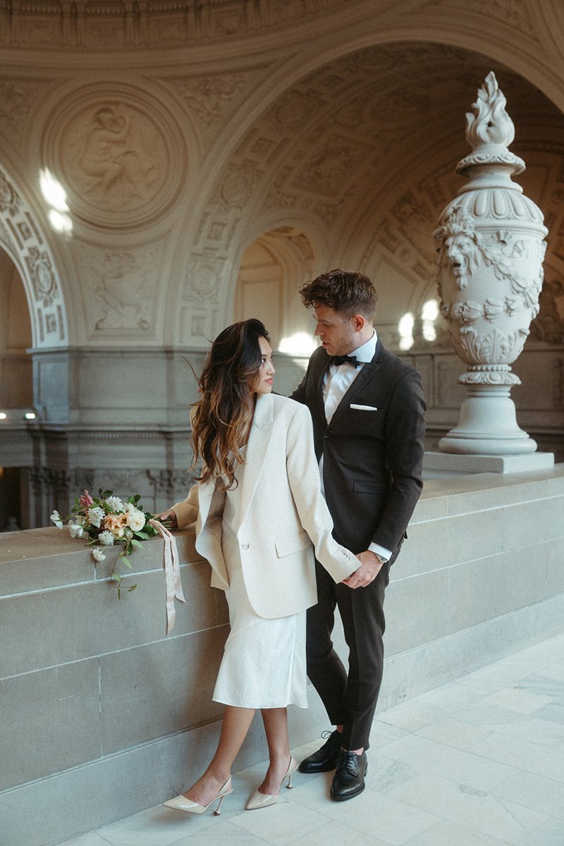 A man and woman in wedding attire standing close in a grand, ornate hall with detailed ceiling carvings. The woman holds a bouquet, and they hold hands while gazing at each other.