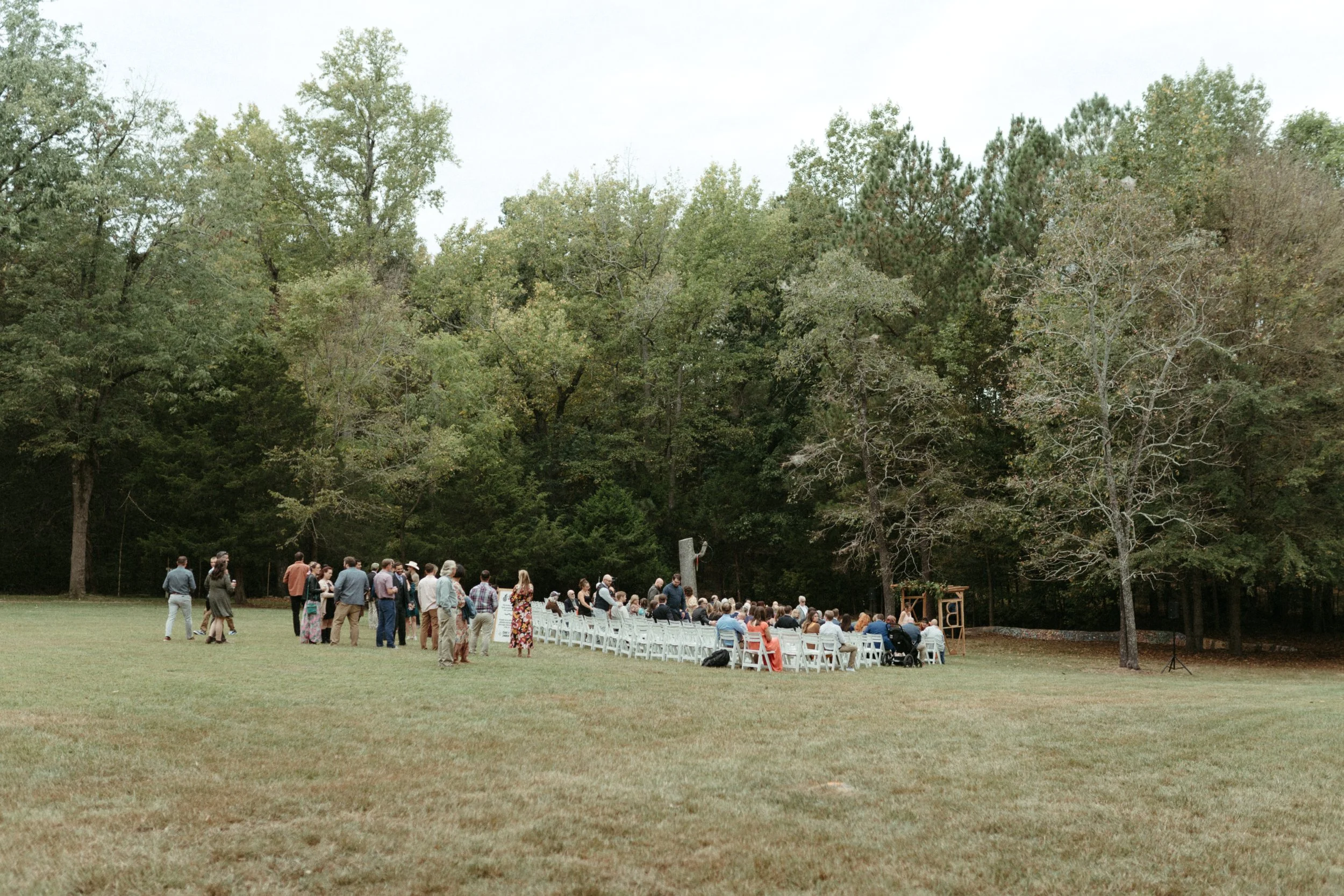 An outdoor wedding ceremony with guests seated in white chairs, surrounded by trees.