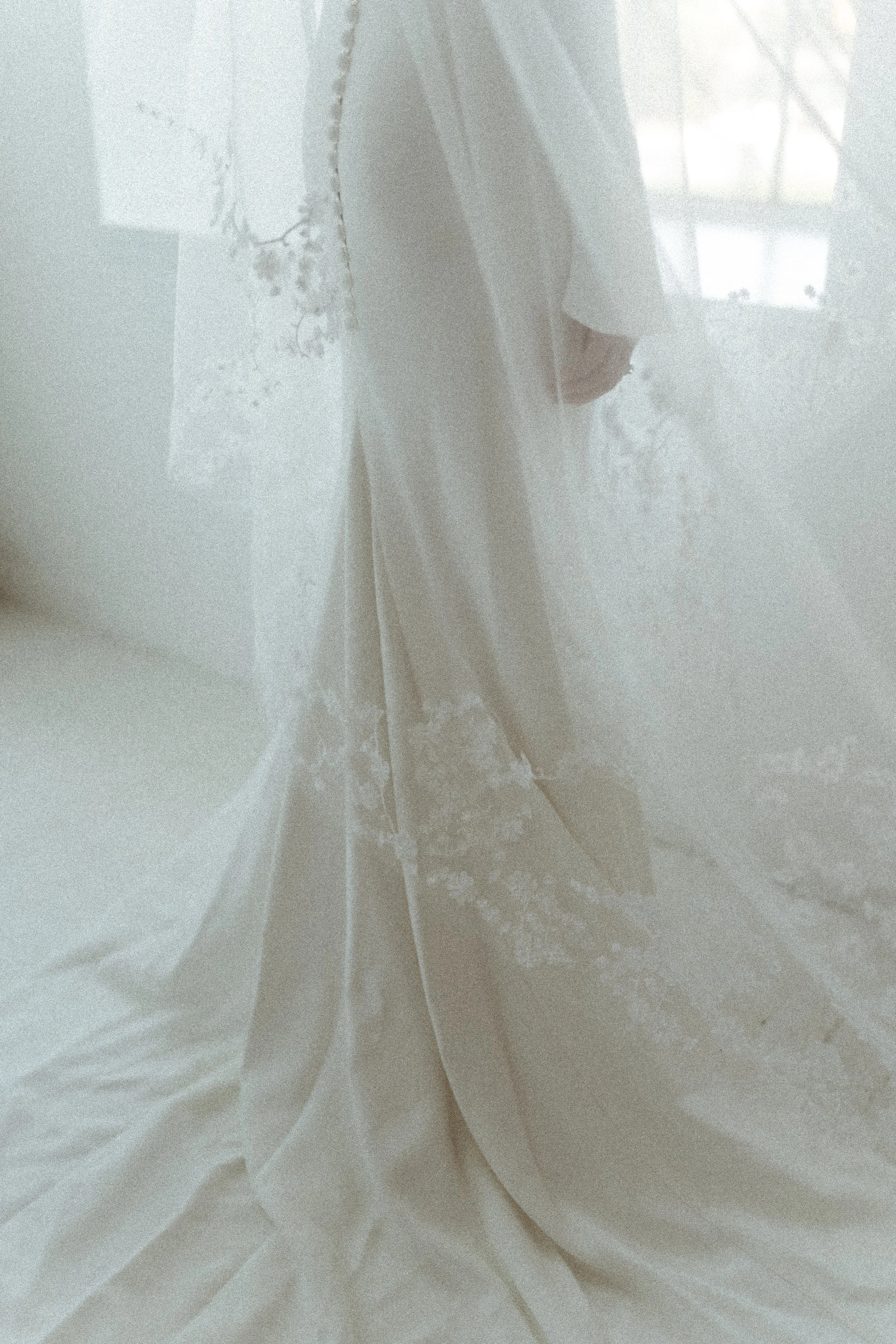 The image shows a person wearing a long, white dress, possibly a wedding gown, seen through a sheer fabric or curtain with lace details. The background includes a window with light coming through.