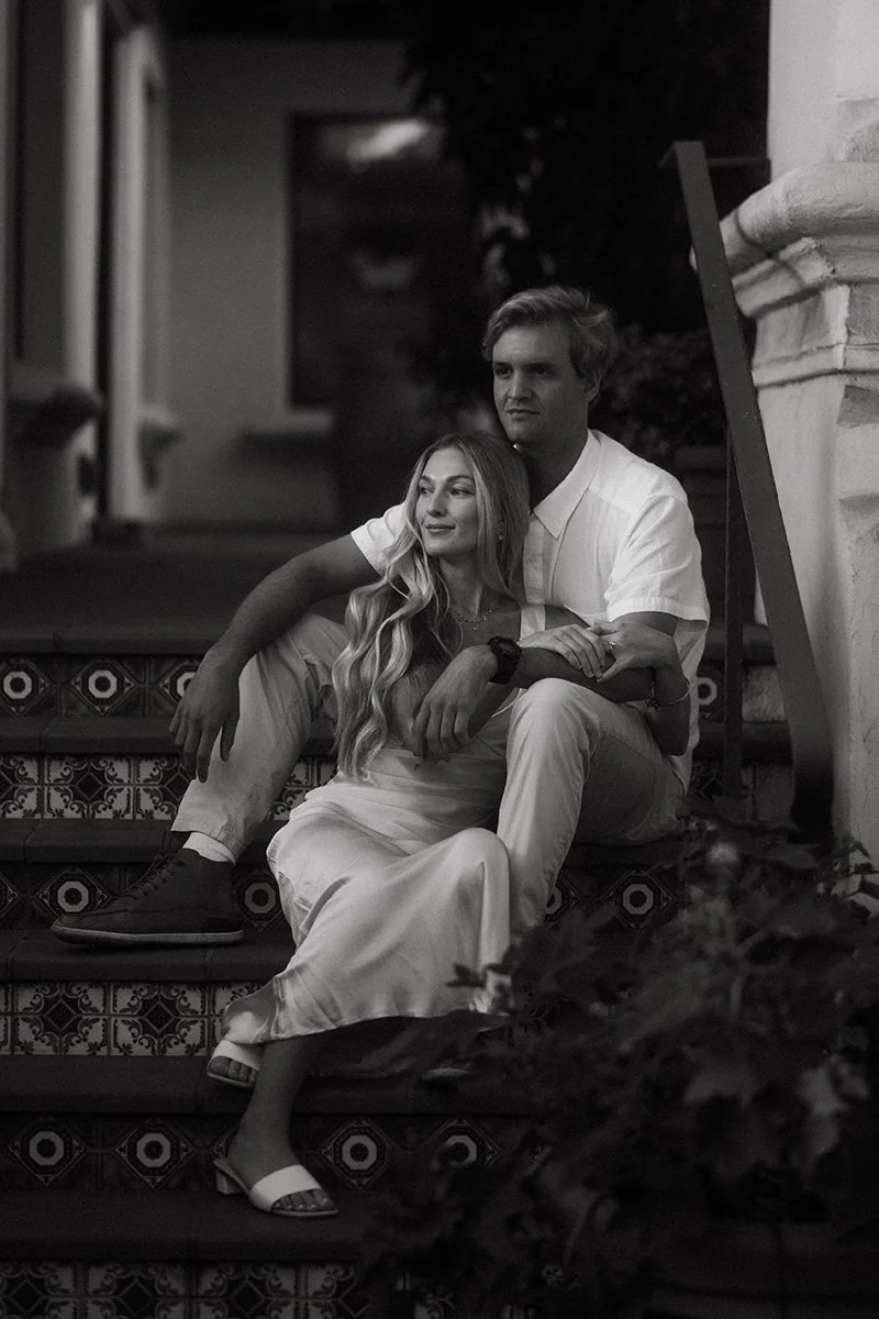 A black and white photo of a young couple sitting on outdoor staircase, with the woman leaning against the man, both looking content, at night.