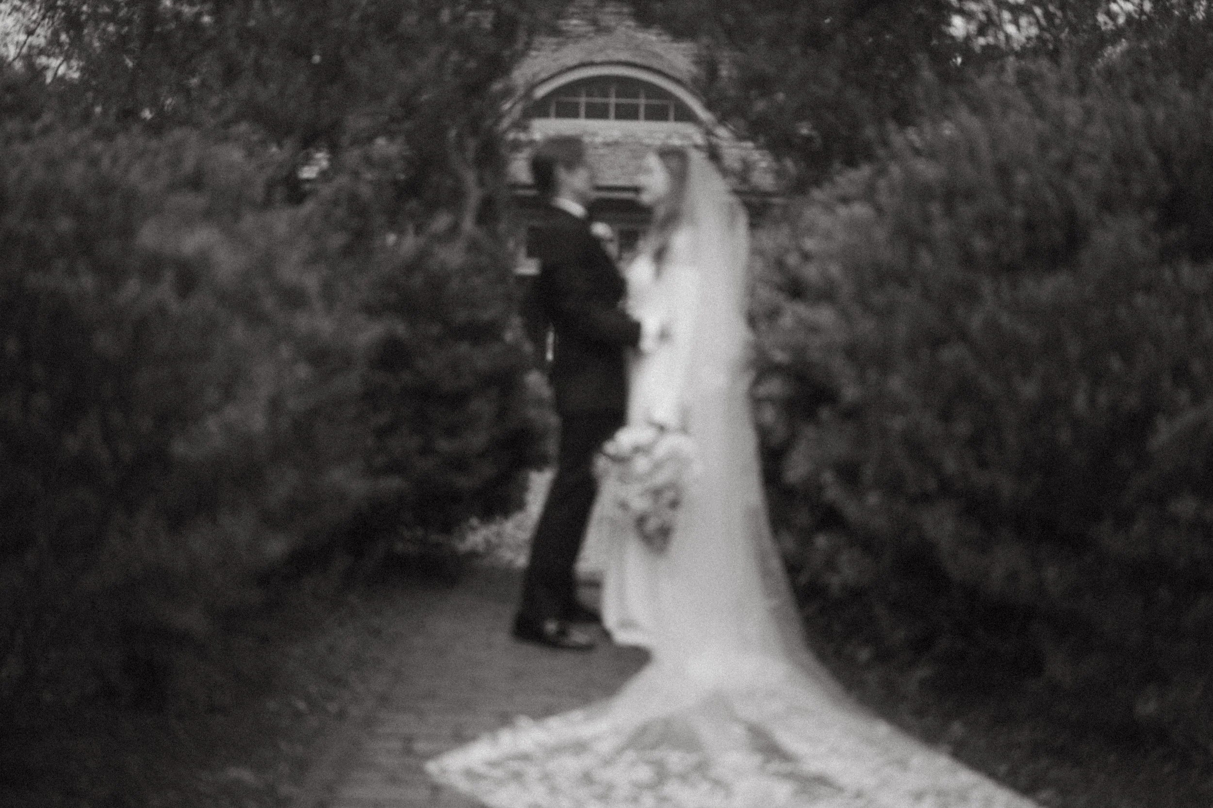 A black and white photo of a bride and groom standing close, holding hands, in an outdoor garden with foliage on both sides and a house in the background.