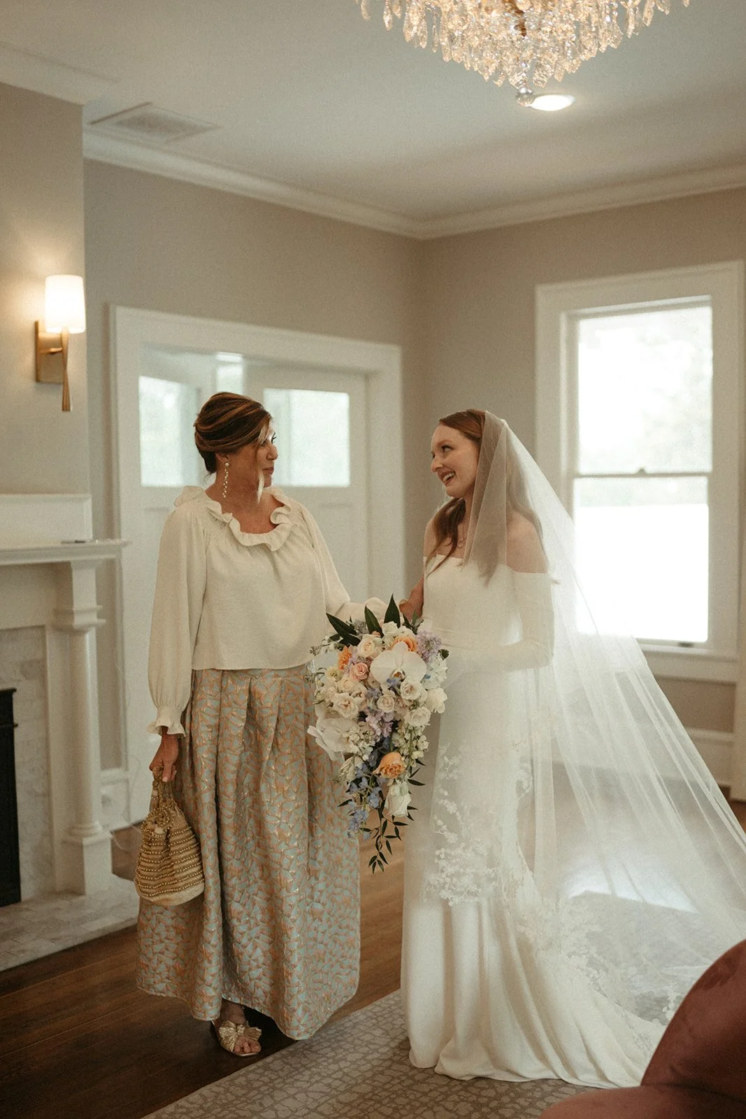 A bride in a white wedding dress and veil holding a bouquet of flowers, smiling and talking to an older woman in a cream blouse and patterned wide-leg pants inside a well-lit room.