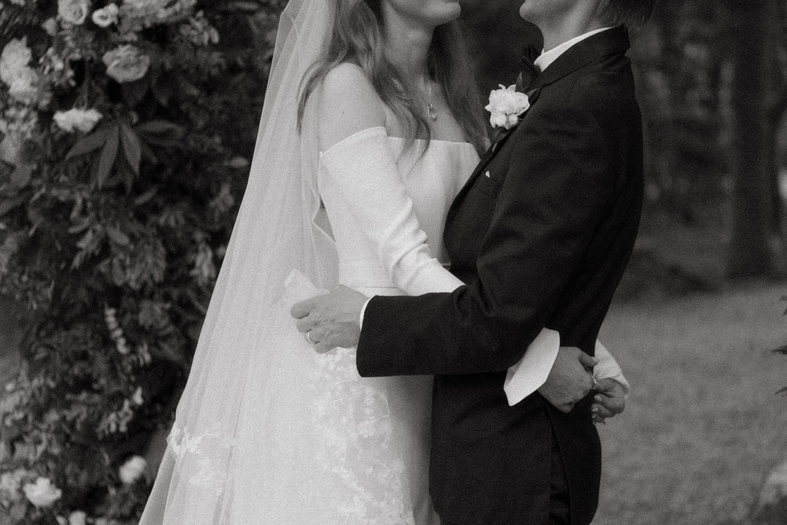 A bride and groom standing close together during their wedding, holding each other's hands, outdoors with foliage in the background.