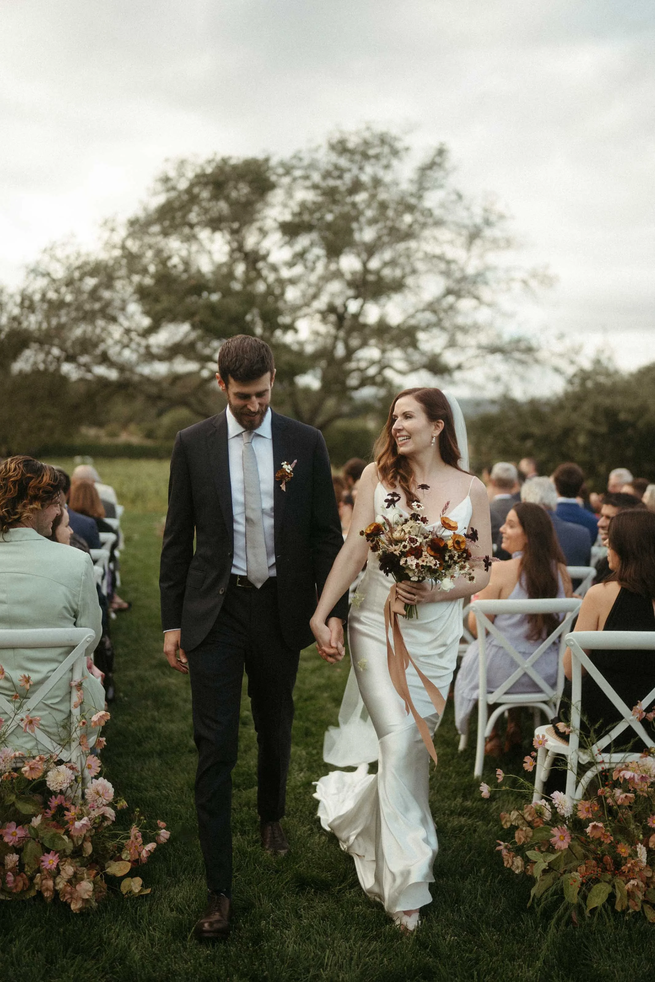 A bride and groom walking hand in hand down the aisle at an outdoor wedding ceremony, surrounded by seated guests, flowers, and trees in the background.