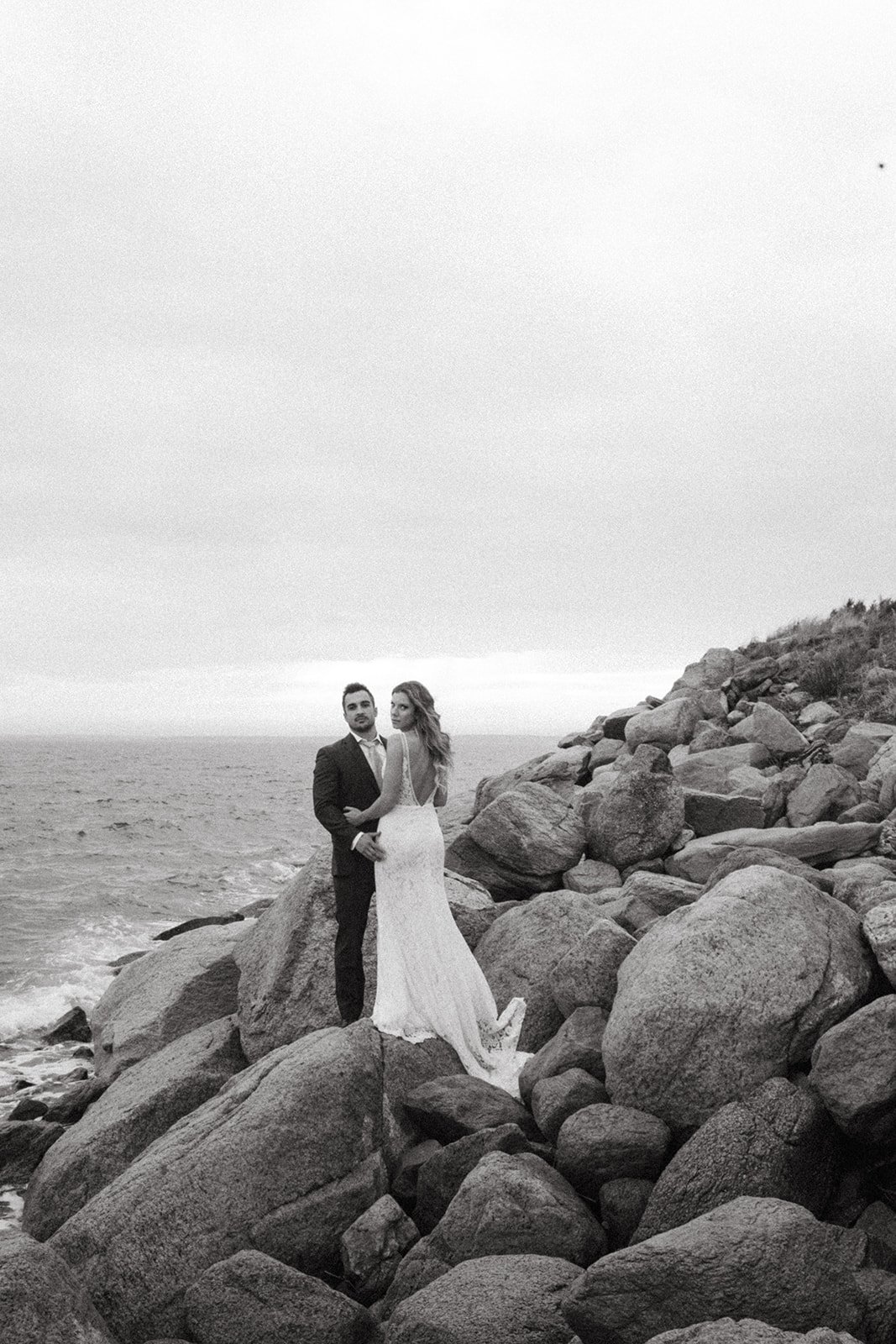 A bride and groom in wedding attire standing on rocks by the ocean.