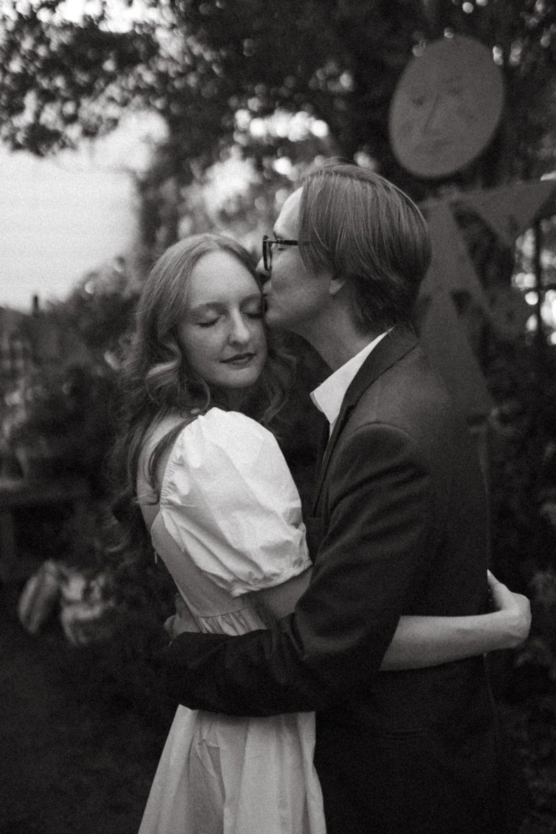 A black and white photo of a couple embracing outdoors. The man is giving the woman a kiss on the forehead, and they are holding each other closely with their eyes closed. There are trees and blurry background scenery.