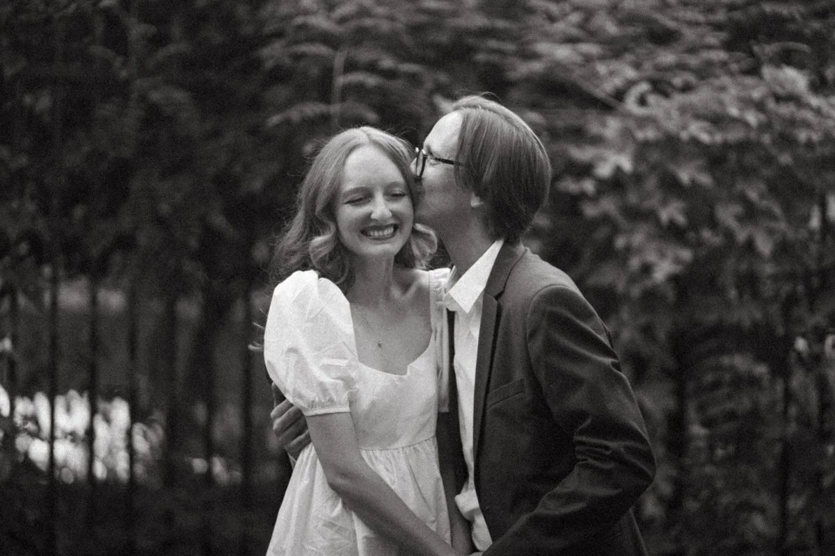 A black-and-white photo of a couple hugging and sharing a kiss on the cheek outdoors, with trees in the background. The woman is smiling, wearing a white dress with puffed sleeves, and the man is wearing glasses and a suit.