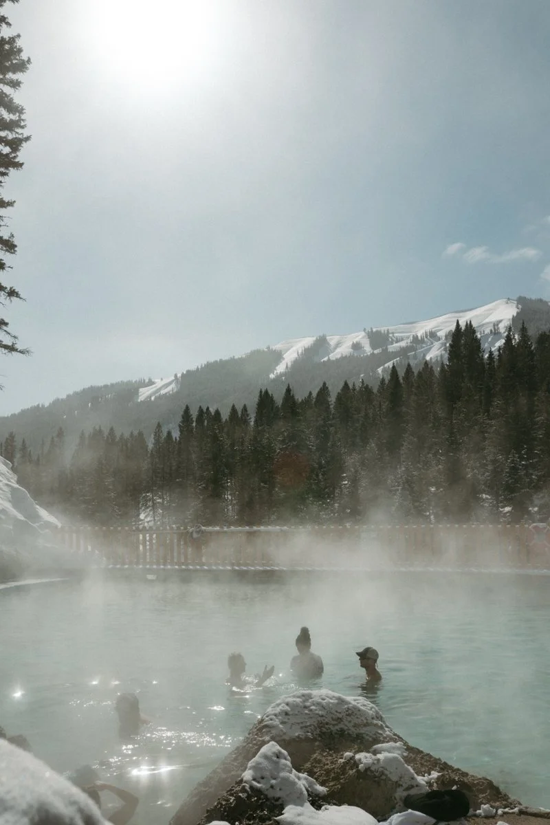 People relaxing in an outdoor hot spring pool with steam rising, surrounded by snow and mountains covered with snow, in a forested area during winter.
