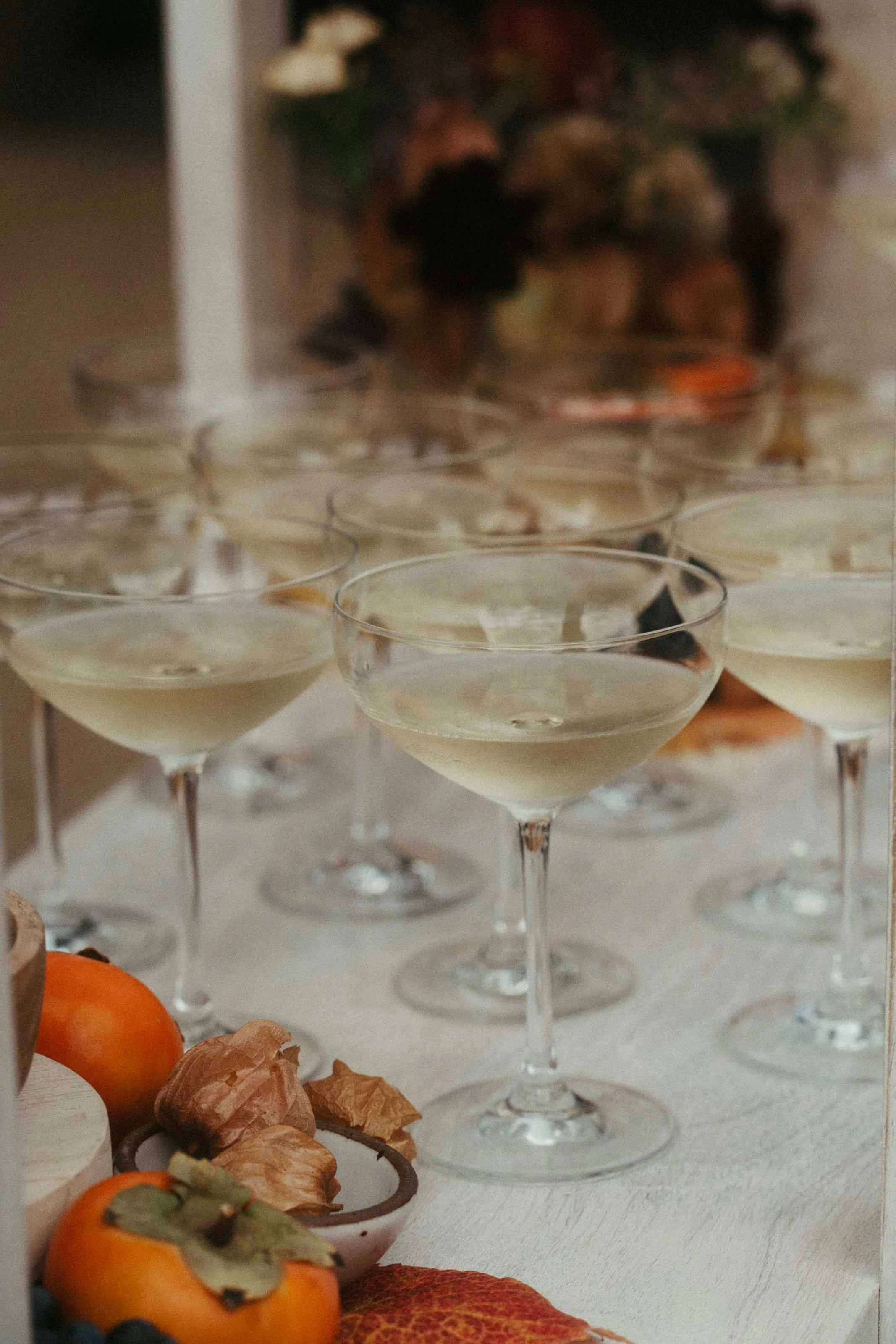 Multiple glasses of champagne or prosecco on a table, with some fruit and autumn leaves in the foreground.