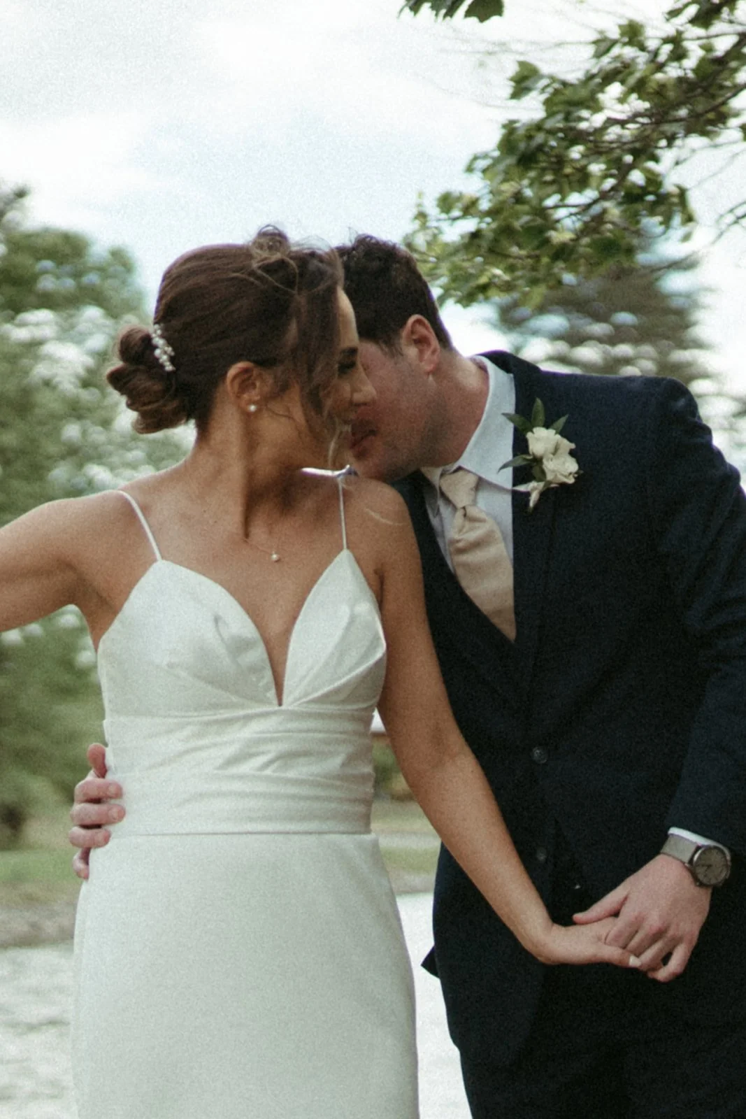 A bride and groom sharing a kiss outdoors near a water body, dressed in wedding attire with trees in the background.