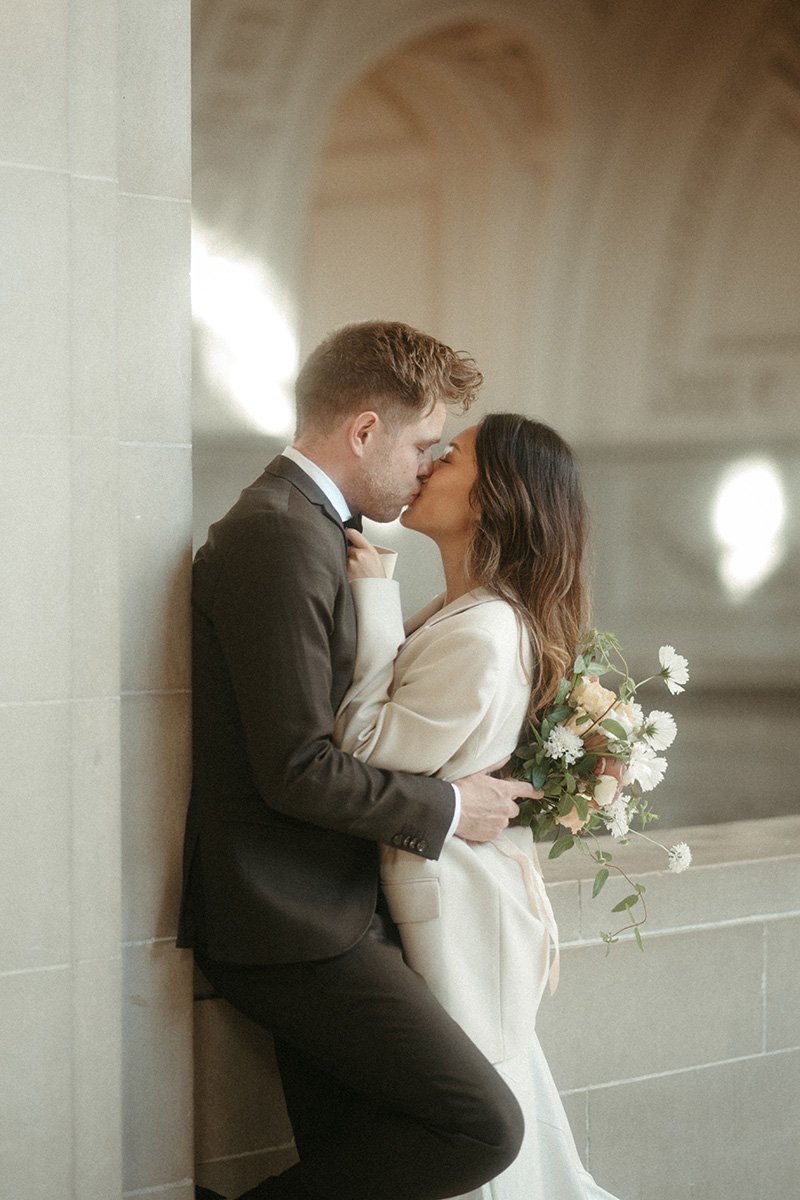 A couple in wedding attire sharing a kiss, with the woman holding a bouquet of white and light pink flowers, while leaning against a beige tiled wall in an indoor setting with blurred architectural details in the background.