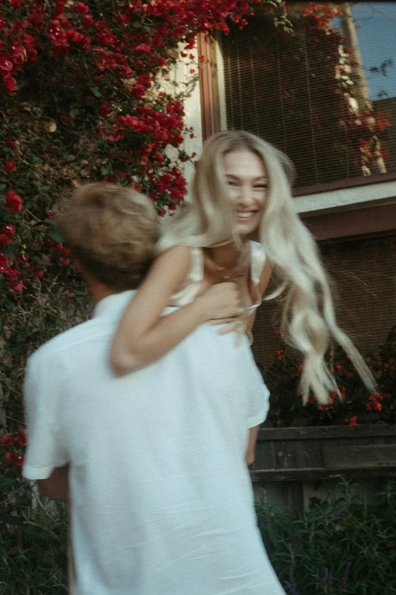 A young woman with long blonde hair smiling as she gets a hug from a young boy in a white shirt, standing outdoors near a house with red flowers and a window with blinds.
