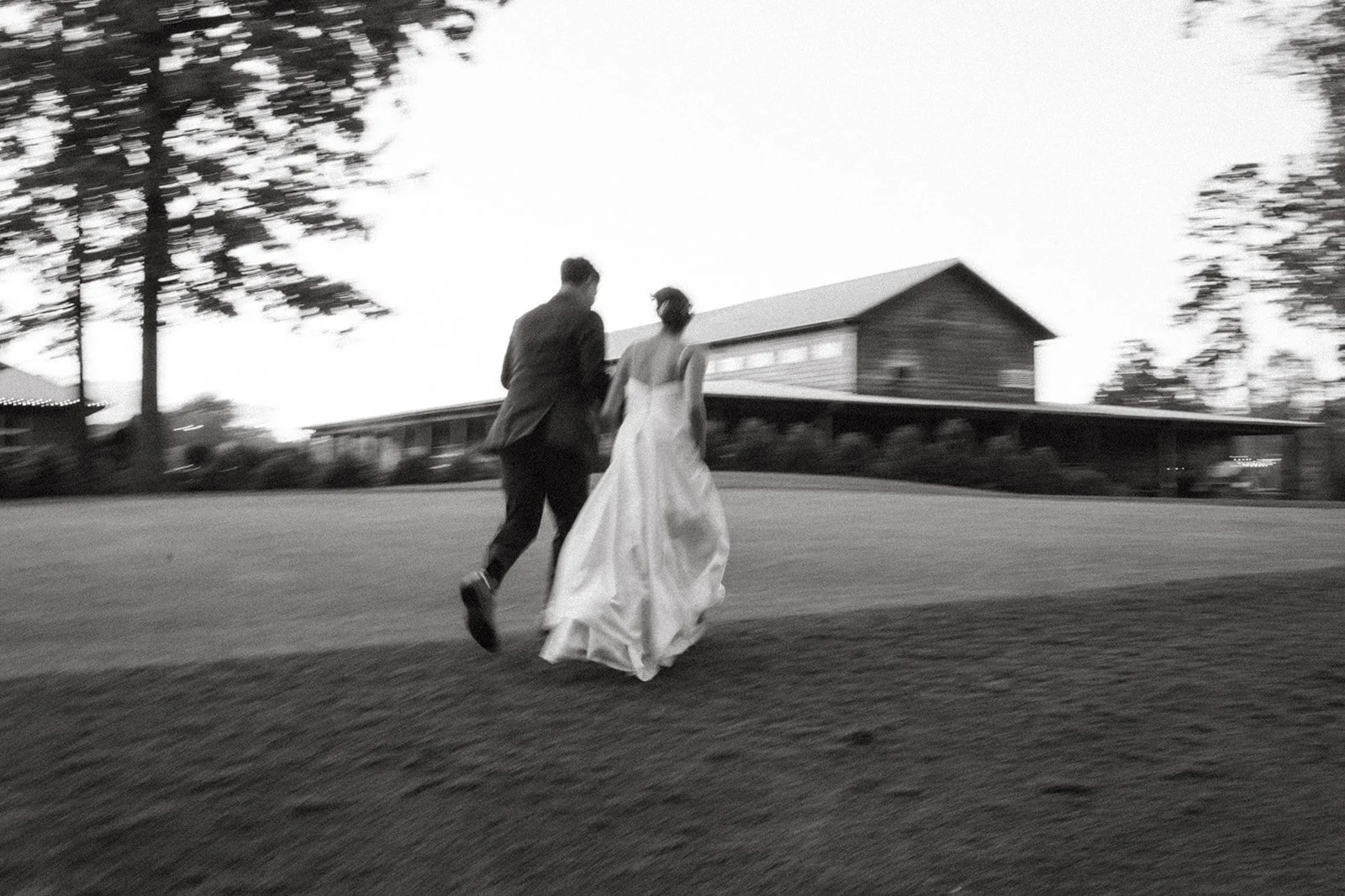 A black and white photo of a couple in wedding attire holding hands and walking outdoors on a slightly sloped grassy area, with trees and a building in the background.