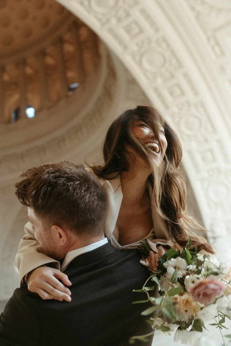 A woman with long brown hair and a white blazer is being lifted in the air by a man in a black suit, holding a bouquet of flowers, inside a grand hall with intricate stone architecture.