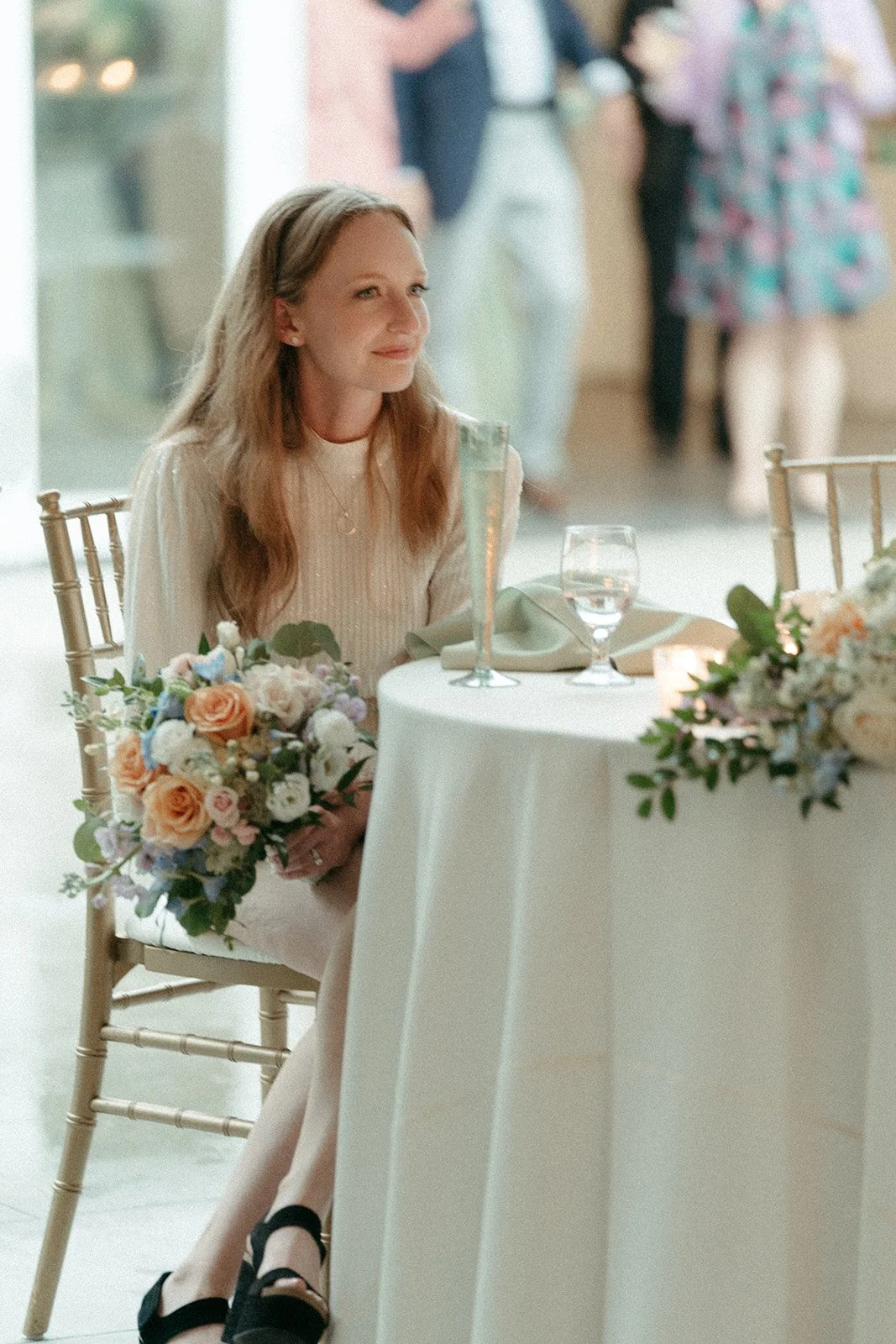 A woman with long hair sitting at a decorated table with a bouquet of flowers, a glass of water, and a champagne flute, during a wedding reception.