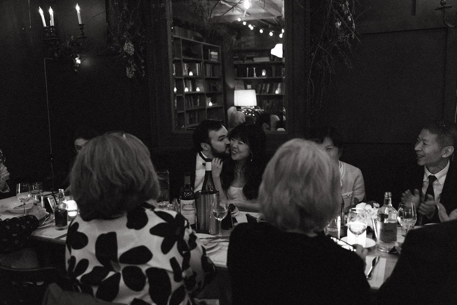 People gathered around a dinner table in a dimly lit restaurant, engaging in conversation, with bottles and glasses on the table, and a bookshelf and mirror in the background.
