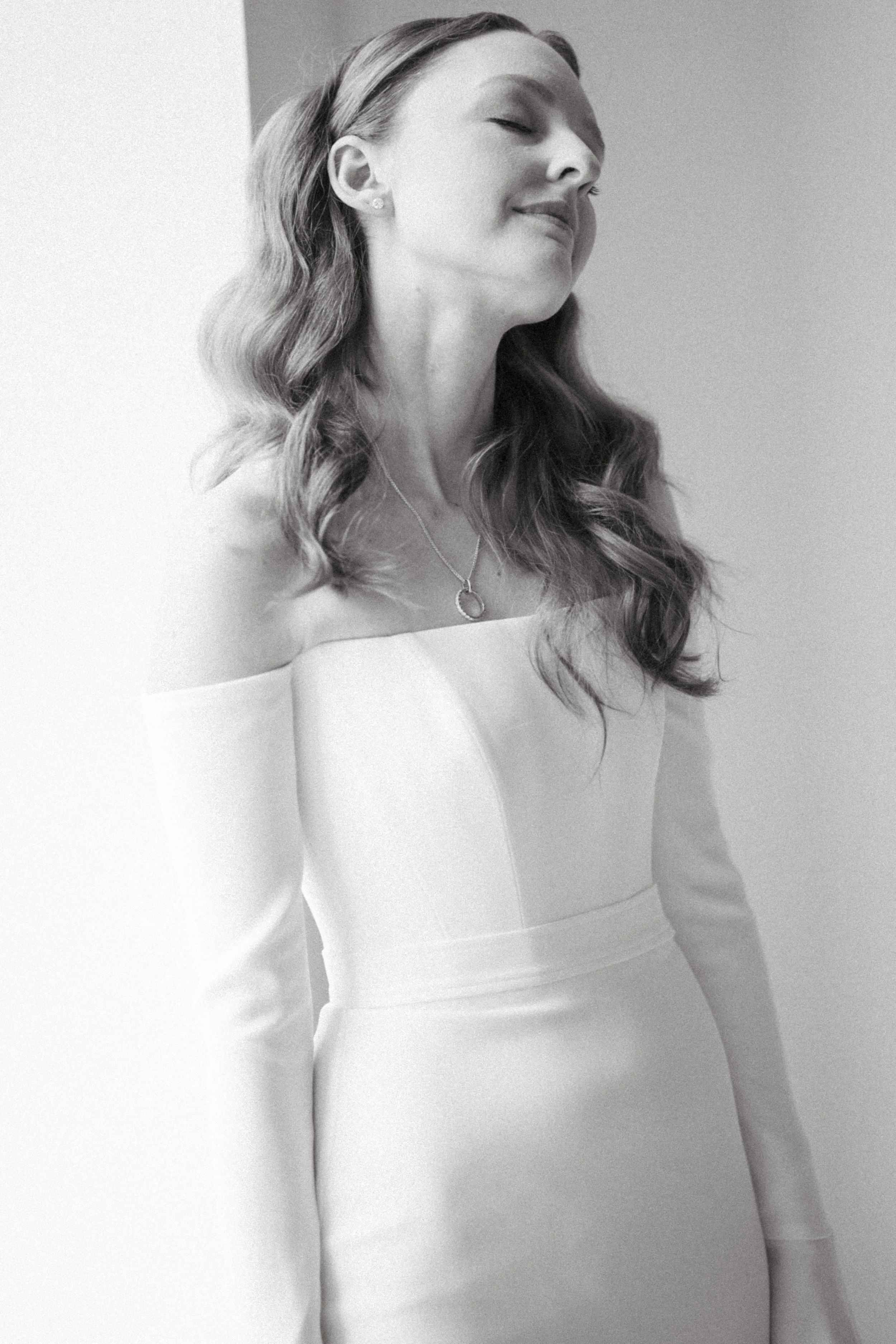 Black and white photo of a woman with long wavy hair wearing a strapless white dress, smiling with her eyes closed.