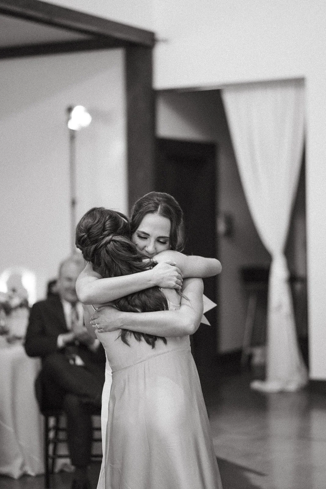 Two women embrace at a wedding, one in a strapless dress, smiling with eyes closed, and the other in a light-colored dress, with dark wavy hair. A man in a suit is seated in the background, clapping.