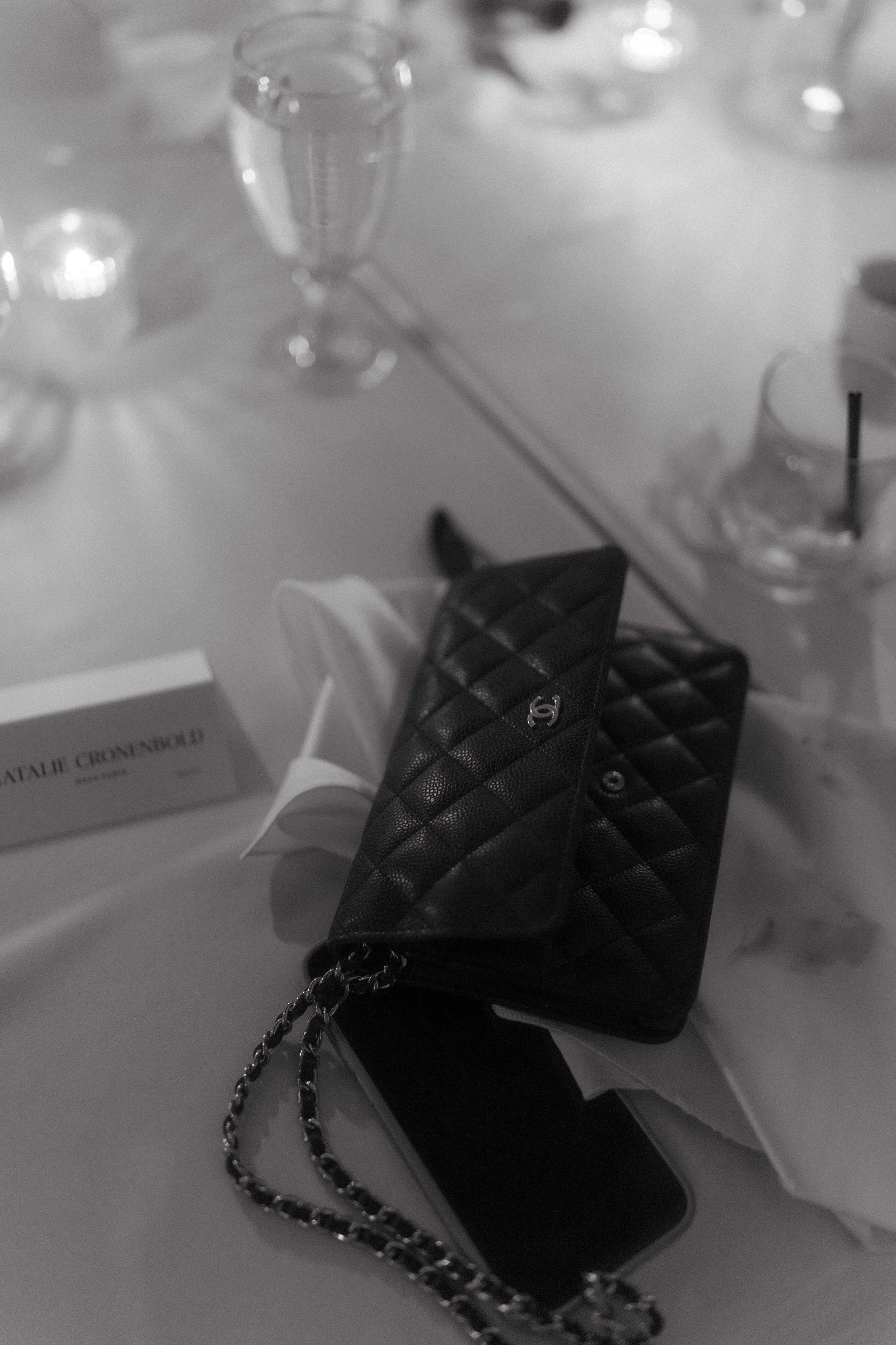 A black quilted Chanel wallet with a chain strap on a table, surrounded by glasses, a box, and a smartphone.