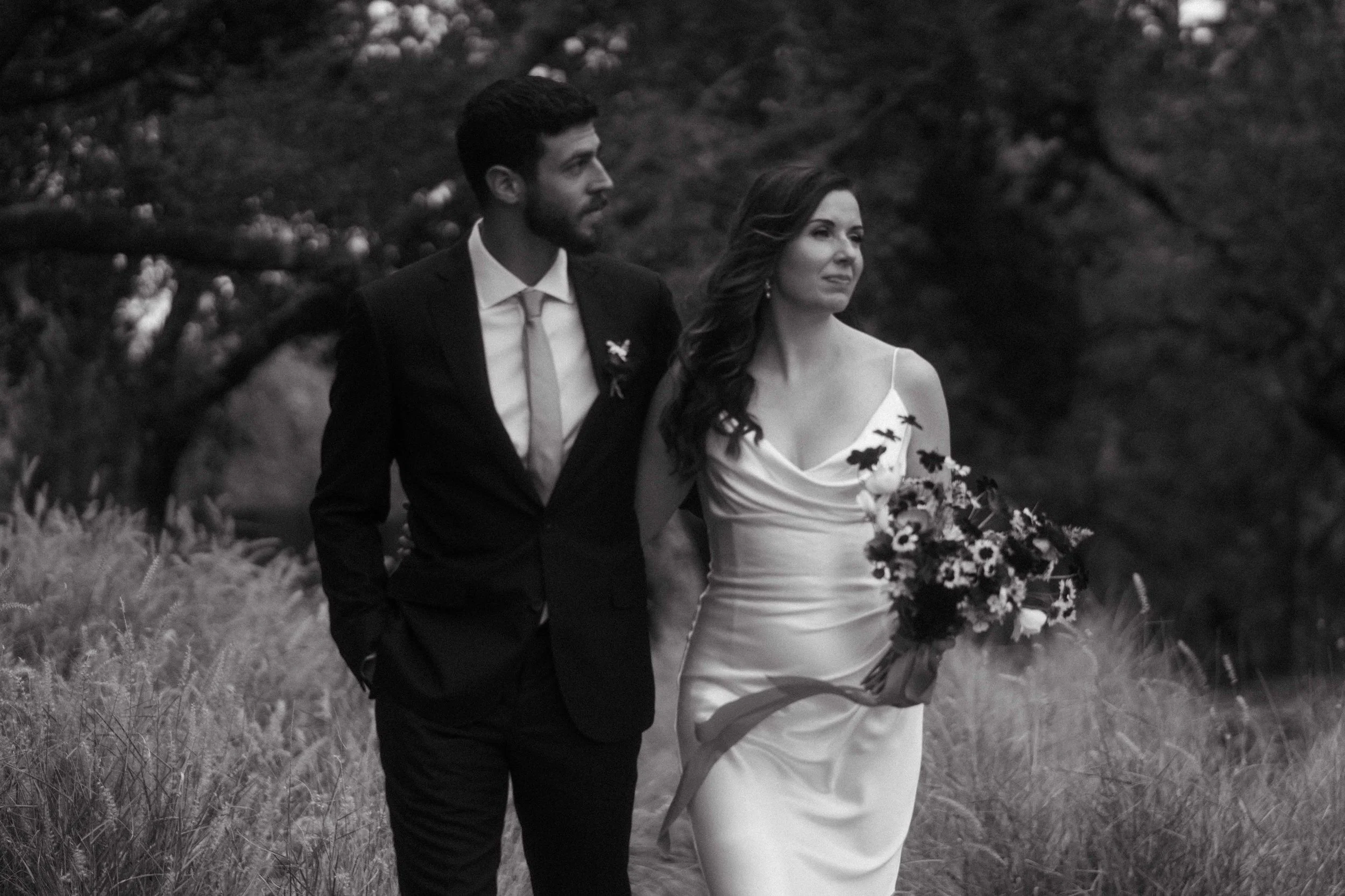 A black and white photo of a bride and groom walking outdoors, with trees and grass in the background. The bride wears a white dress and holds a bouquet, and the groom is in a dark suit and tie.