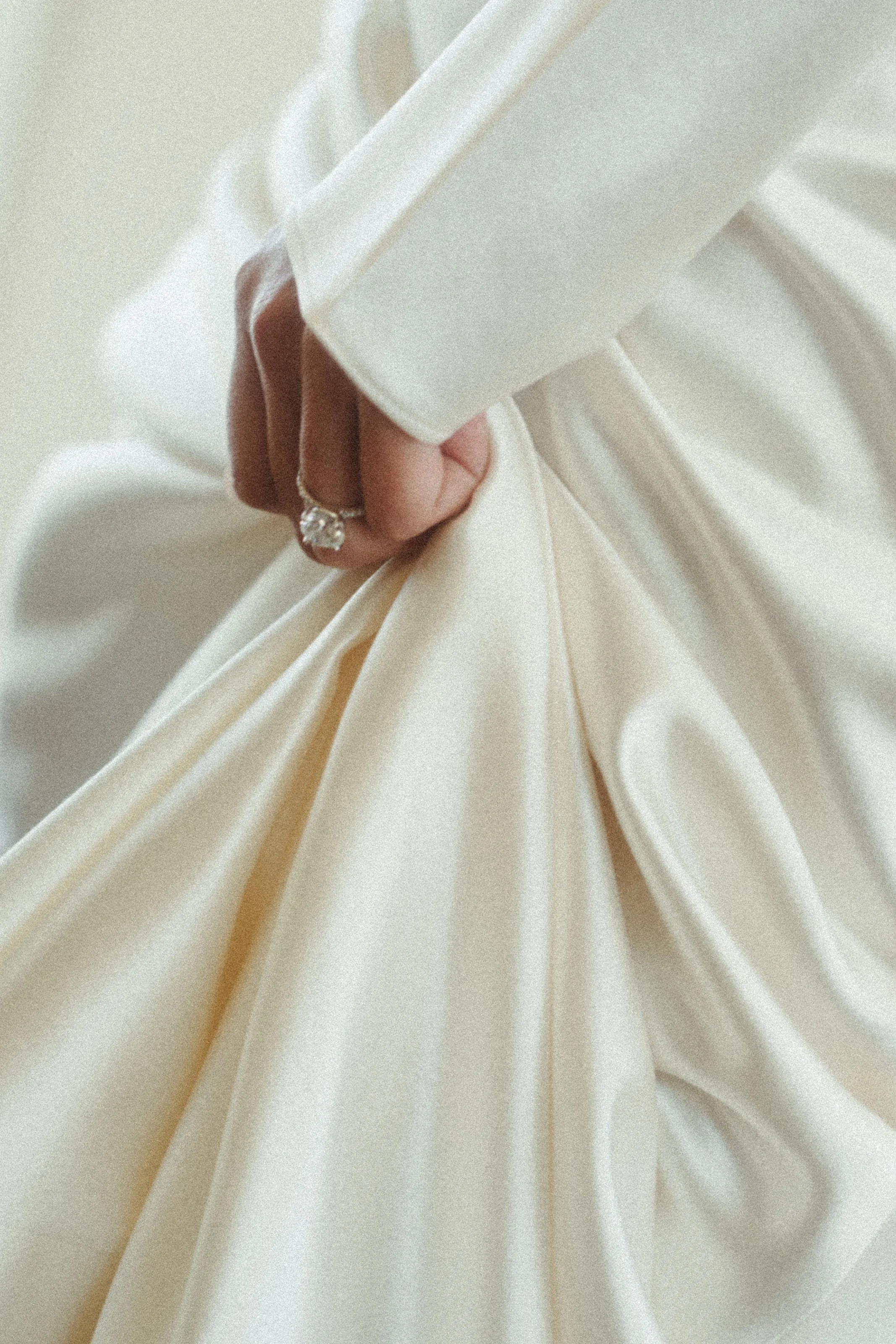 Close-up of a hand holding the fabric of a cream-colored satin wedding dress, wearing a large diamond engagement ring.