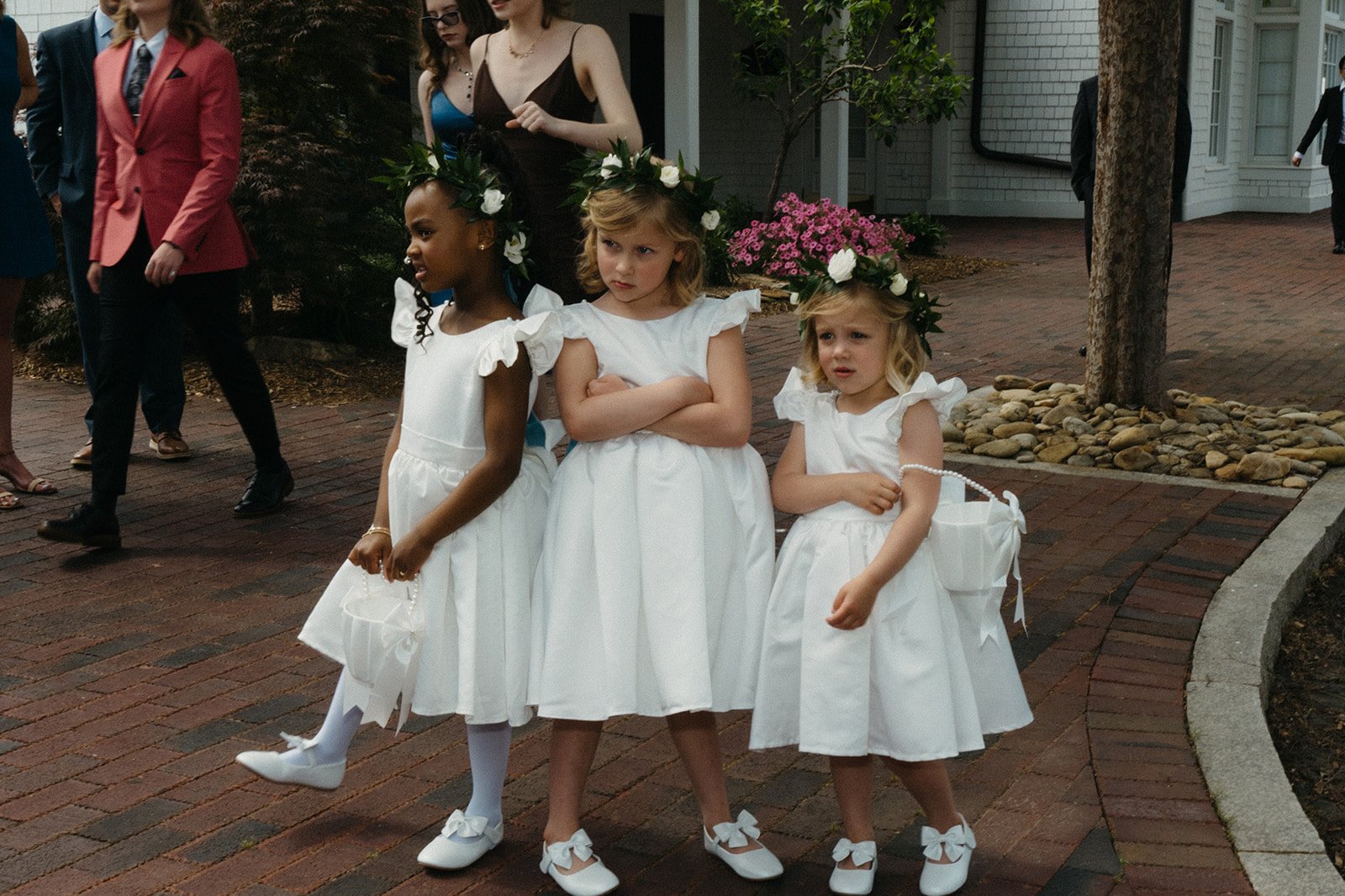Three young girls dressed in white dresses and floral crowns are standing on a brick sidewalk during a wedding or formal event. One girl is crossing her leg, a middle girl has arms crossed, and the third girl holds a small bag, all wearing matching w