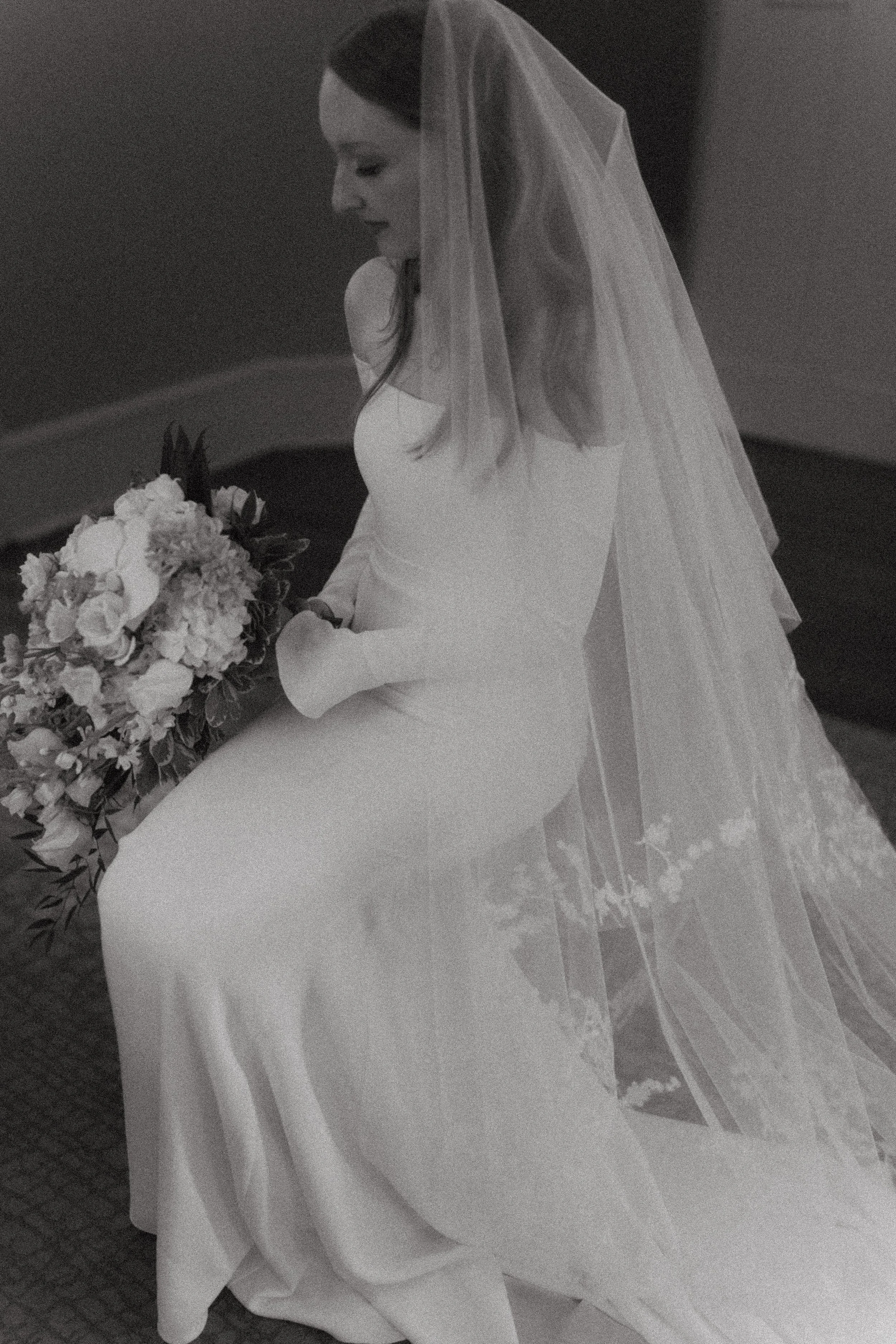 A bride in a wedding dress and veil sitting on a bench and holding a bouquet of flowers.