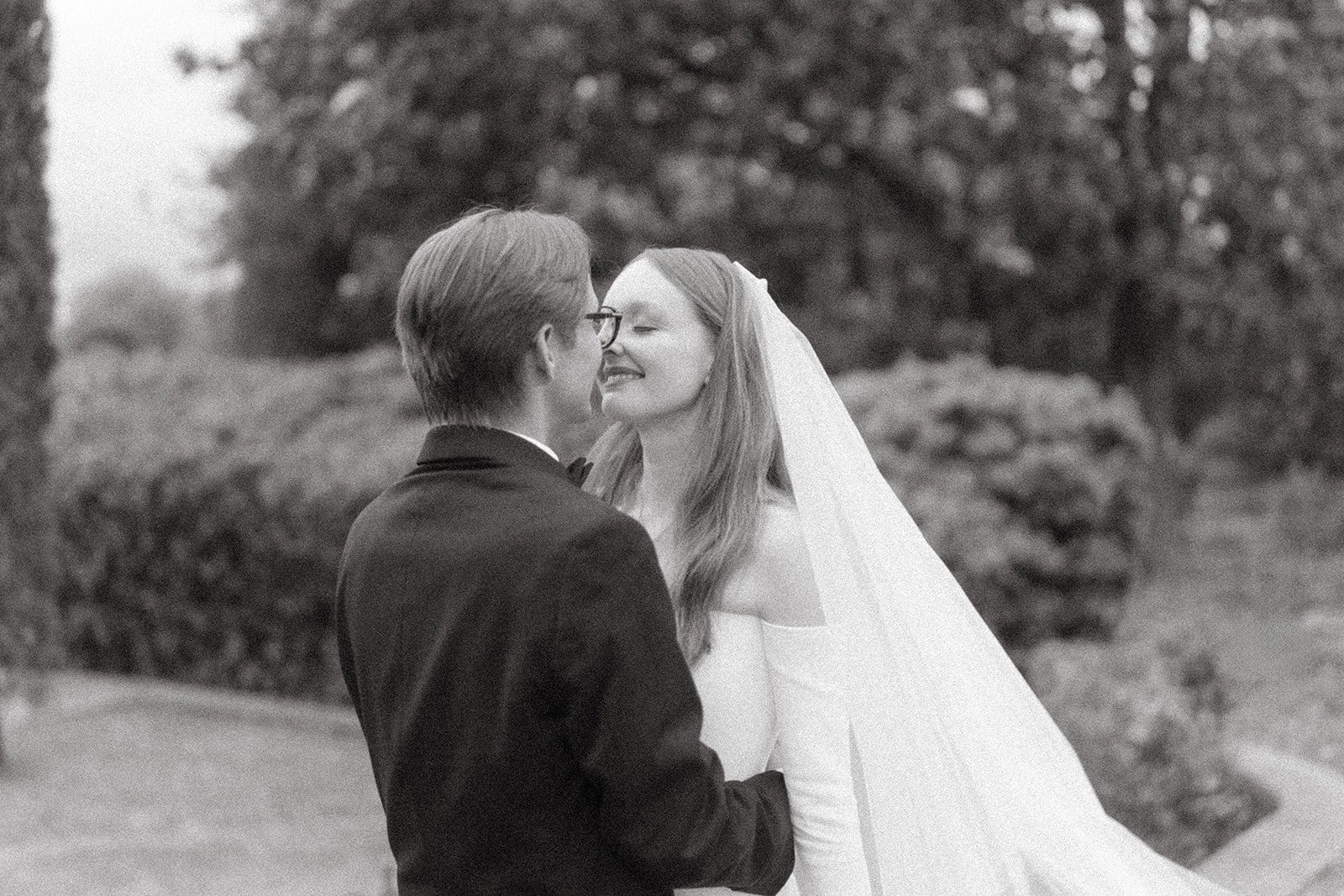 A black and white photo of a bride and groom smiling at each other, standing outdoors with trees in the background. The bride has long hair, a veil, and is wearing an off-the-shoulder wedding gown. The groom is wearing glasses and a suit.