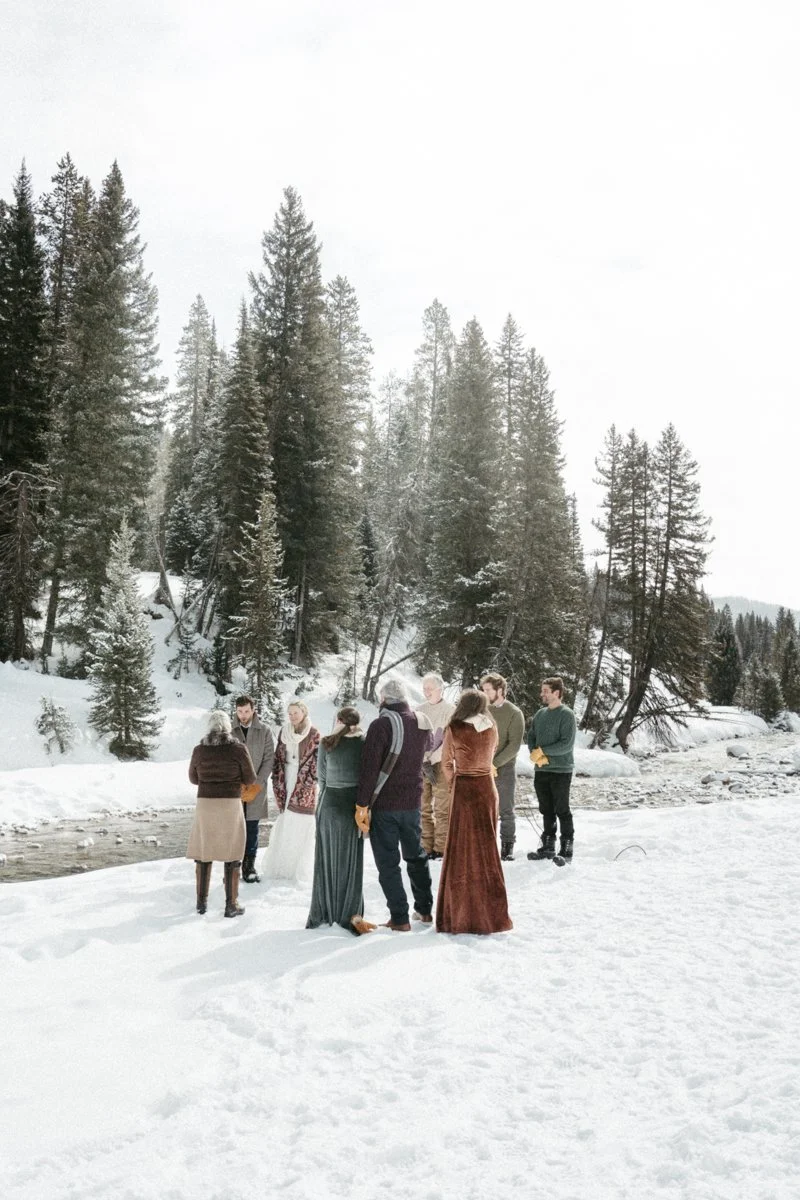 Group of people standing on snow near a river in a snowy forest with tall pine trees.