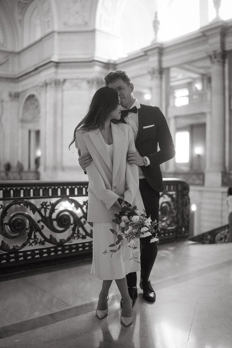 A couple dressed in formal attire in an elegant, ornate hall. The woman is holding a bouquet of flowers, and the man has his arm around her.