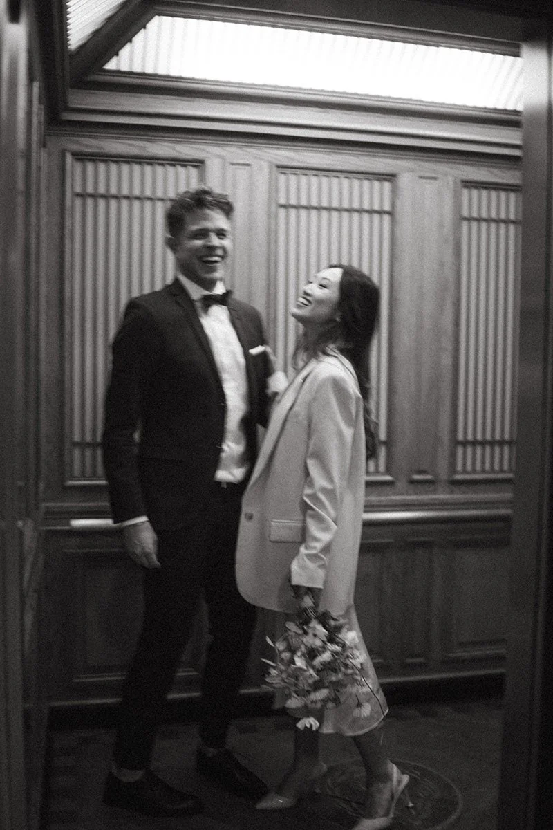 A black-and-white photo of a man in a tuxedo and a woman in a blazer and floral skirt, both smiling and looking happy together. The woman is holding a bouquet of flowers.