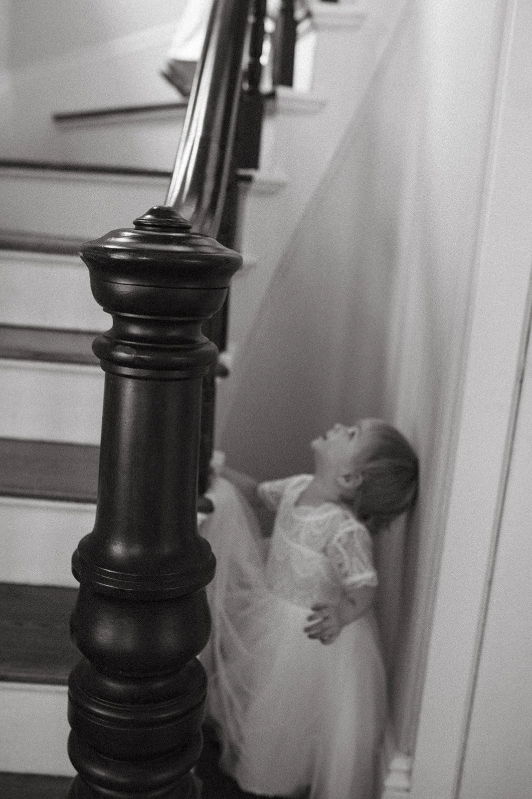 A young girl wearing a lace dress arching her back under a staircase railing, laughing or smiling.