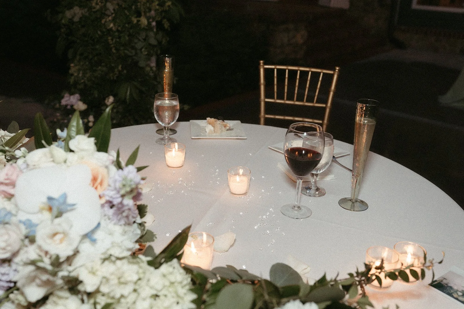 A round table set for a romantic dinner with floral centerpiece, candles, and glasses of water, wine, and champagne at night.