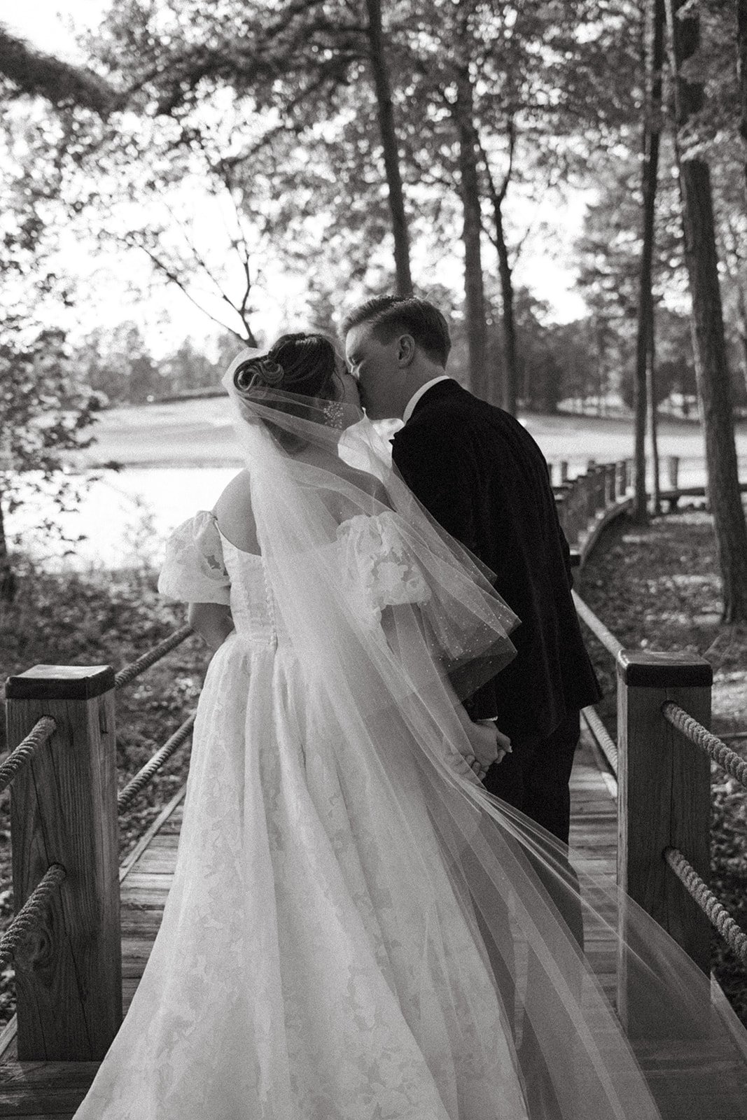A bride and groom share a kiss on a wooden bridge near a lake or pond, surrounded by trees, in black and white.