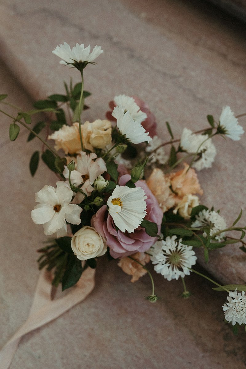 A bouquet of various flowers including white, pink, and cream-colored blossoms with green leaves, resting on a beige surface.