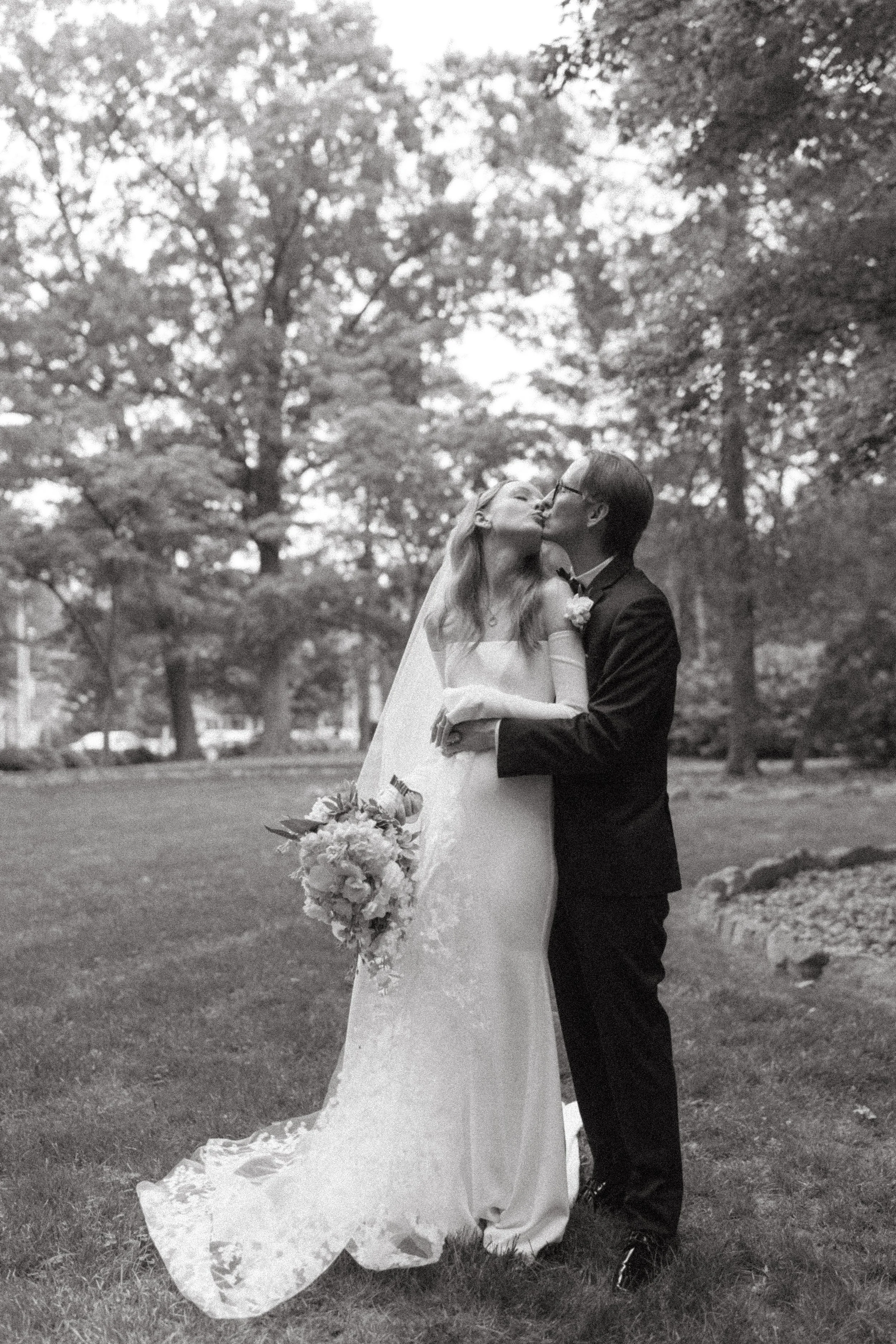 A bride and groom kissing outdoors on their wedding day, with trees in the background, the bride holding a bouquet, in black and white.