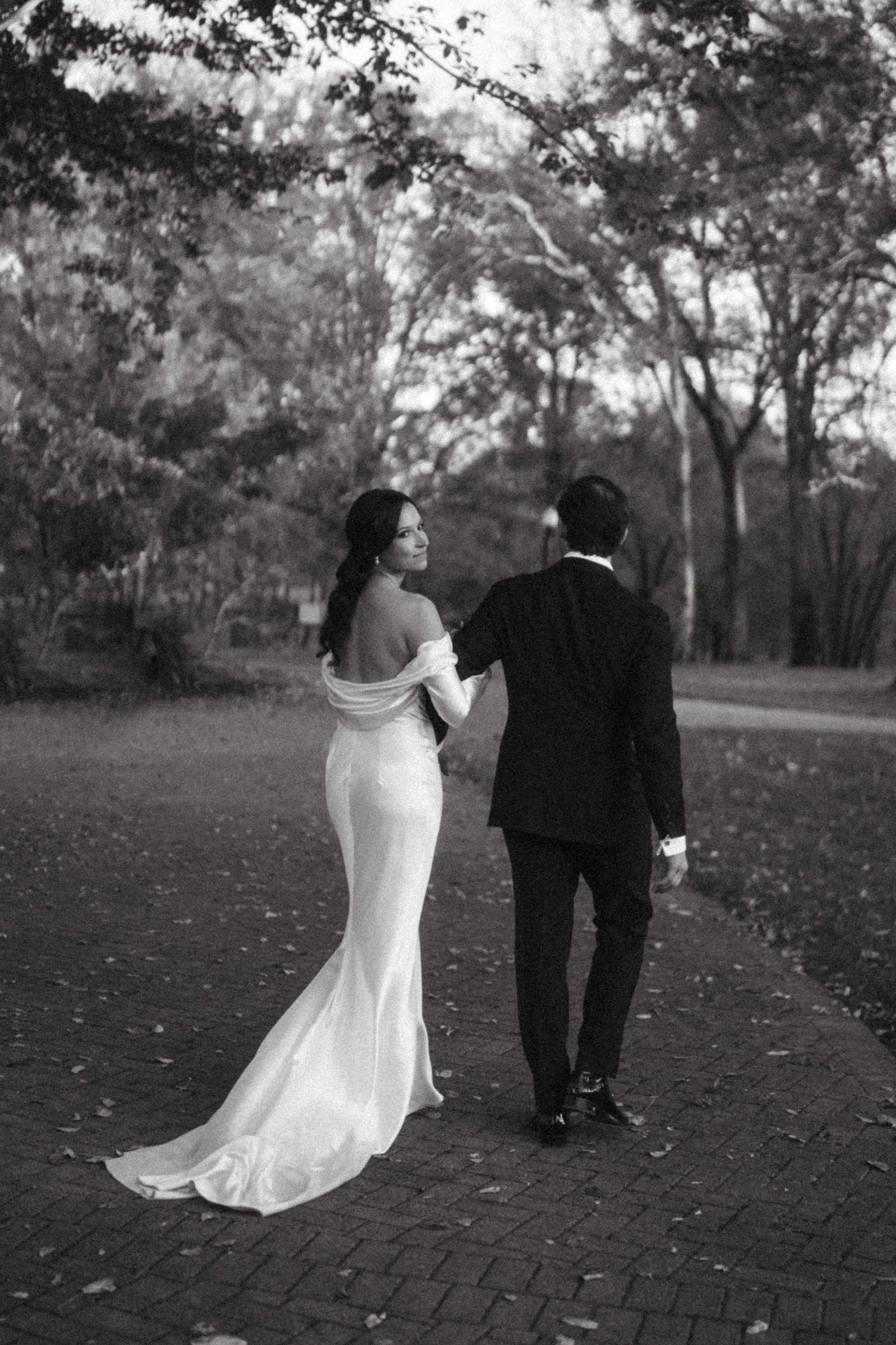 A couple walking hand in hand through a park during twilight. The woman is wearing a long, elegant, off-shoulder white wedding gown, and the man is dressed in a black tuxedo. The background features trees with sparse leaves, and the ground is paved w