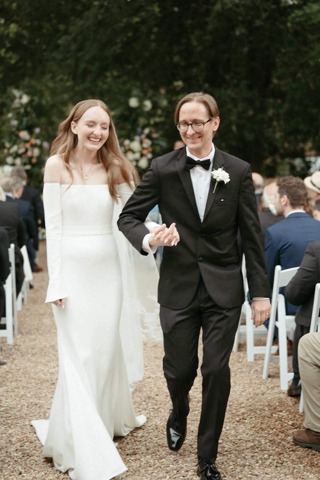 A bride and groom walking down the aisle at an outdoor wedding ceremony, smiling and holding hands, with guests seated on either side.