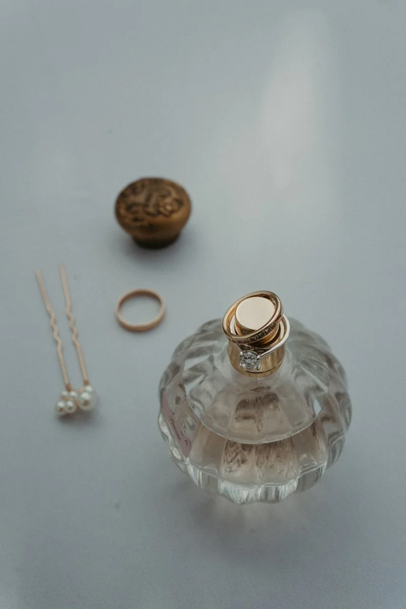 Jewelry and perfume bottle on a white surface, including a gold ring, pearl earrings, a gold ring, a ring with a gemstone, and a small container with a cork stopper.