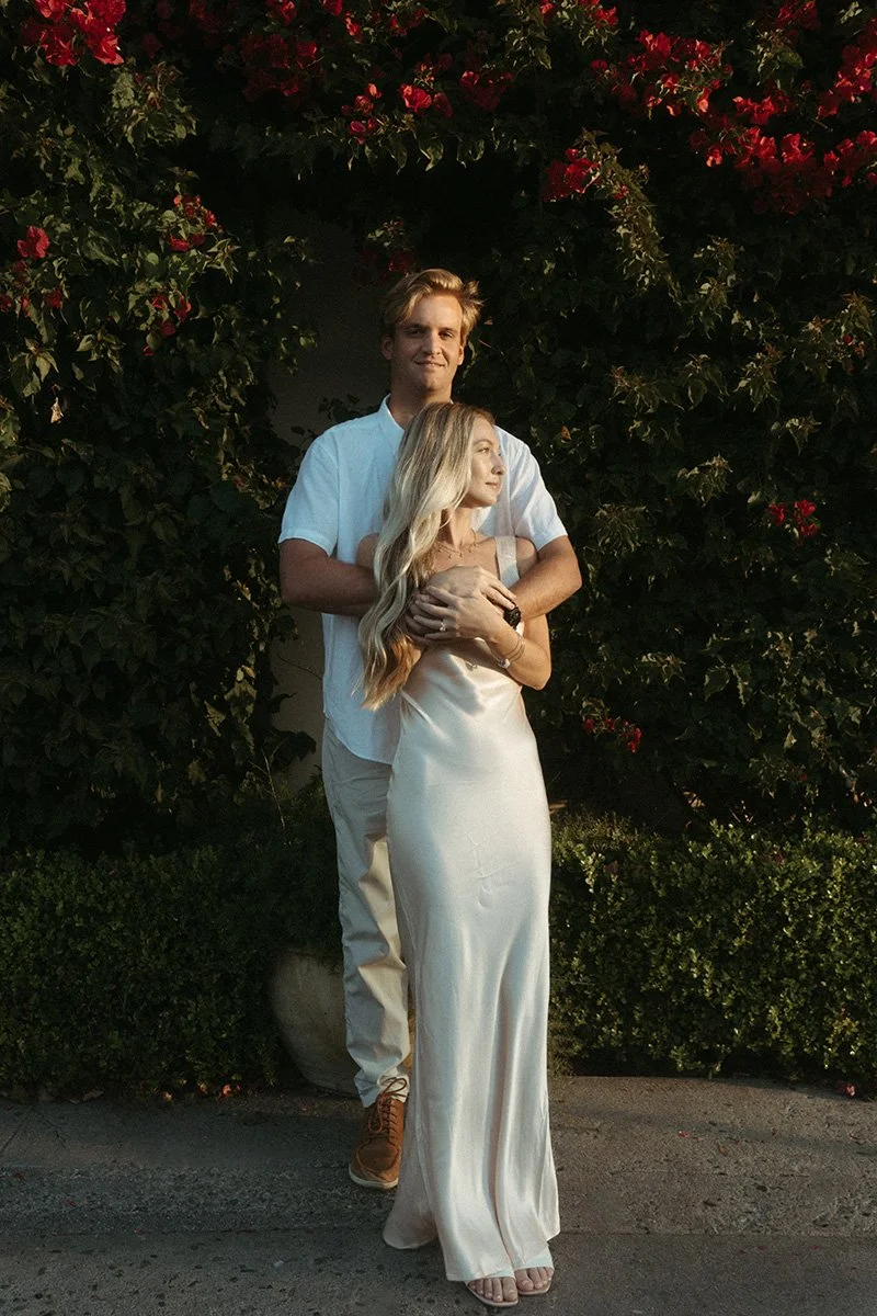 A young man and woman standing together outdoors near flowering bushes at dusk, with the man embracing the woman from behind.