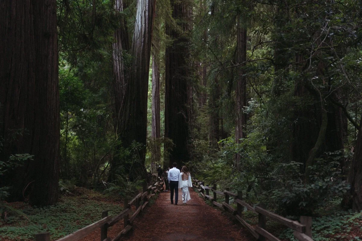 Couple walking hand in hand on a forest trail surrounded by tall trees and lush greenery.