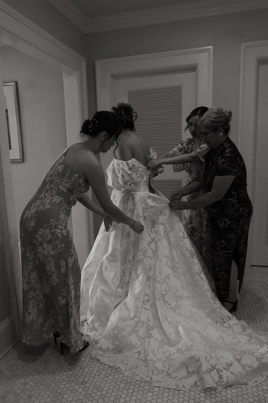 a black and white image of a bride's mom and sister helping her zip her wedding dress