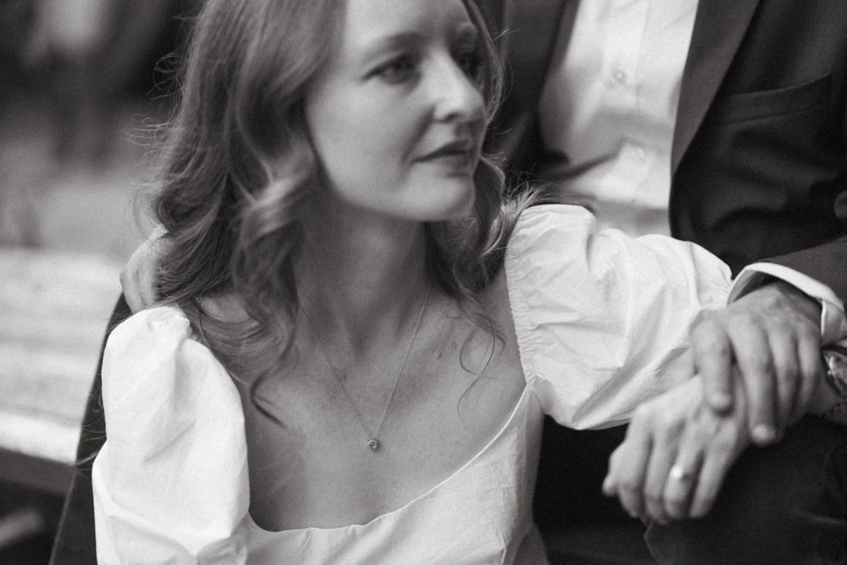 A woman with wavy hair wearing a white blouse and a necklace, sitting and holding hands with a man in a suit.