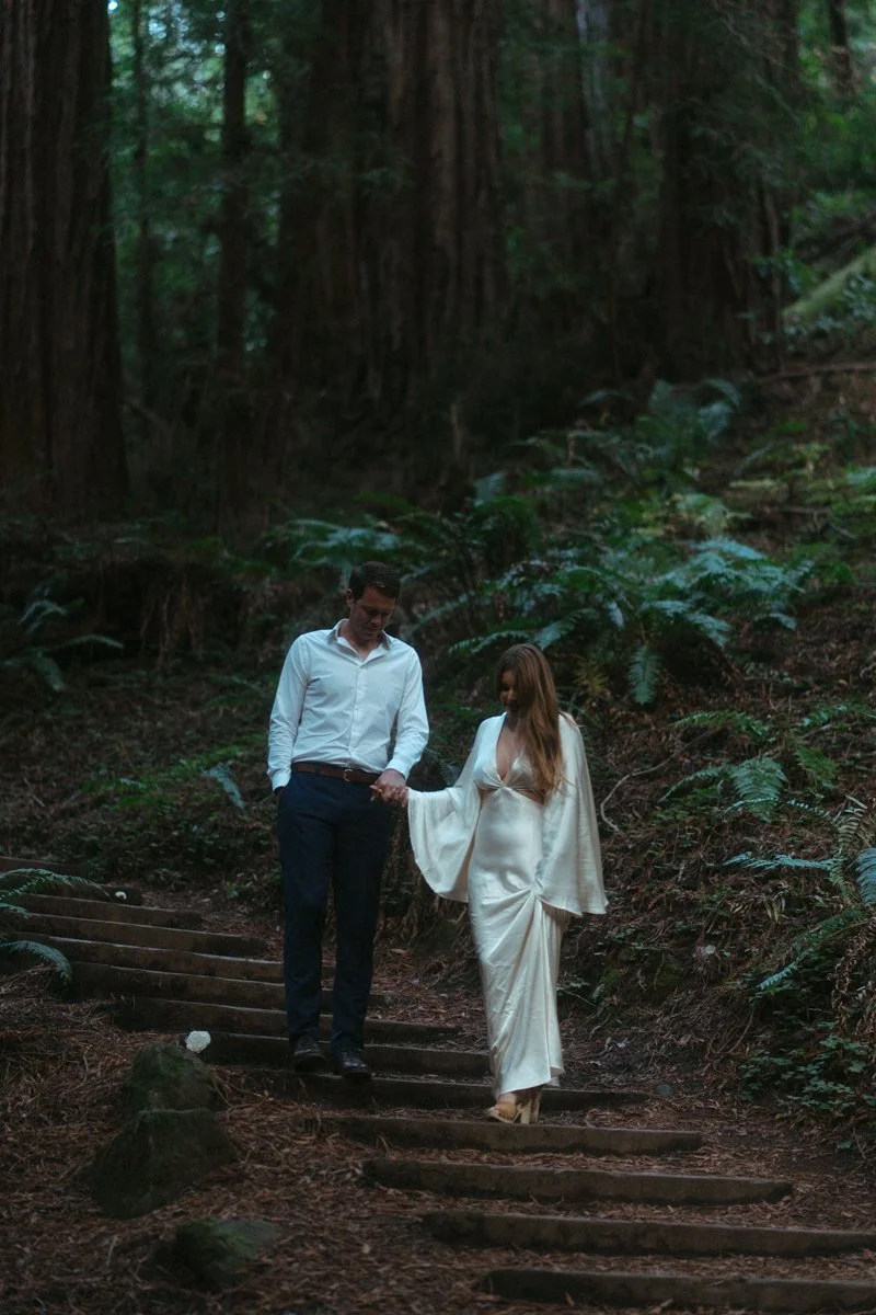 A couple holding hands walking down a forest trail surrounded by tall trees and lush greenery.